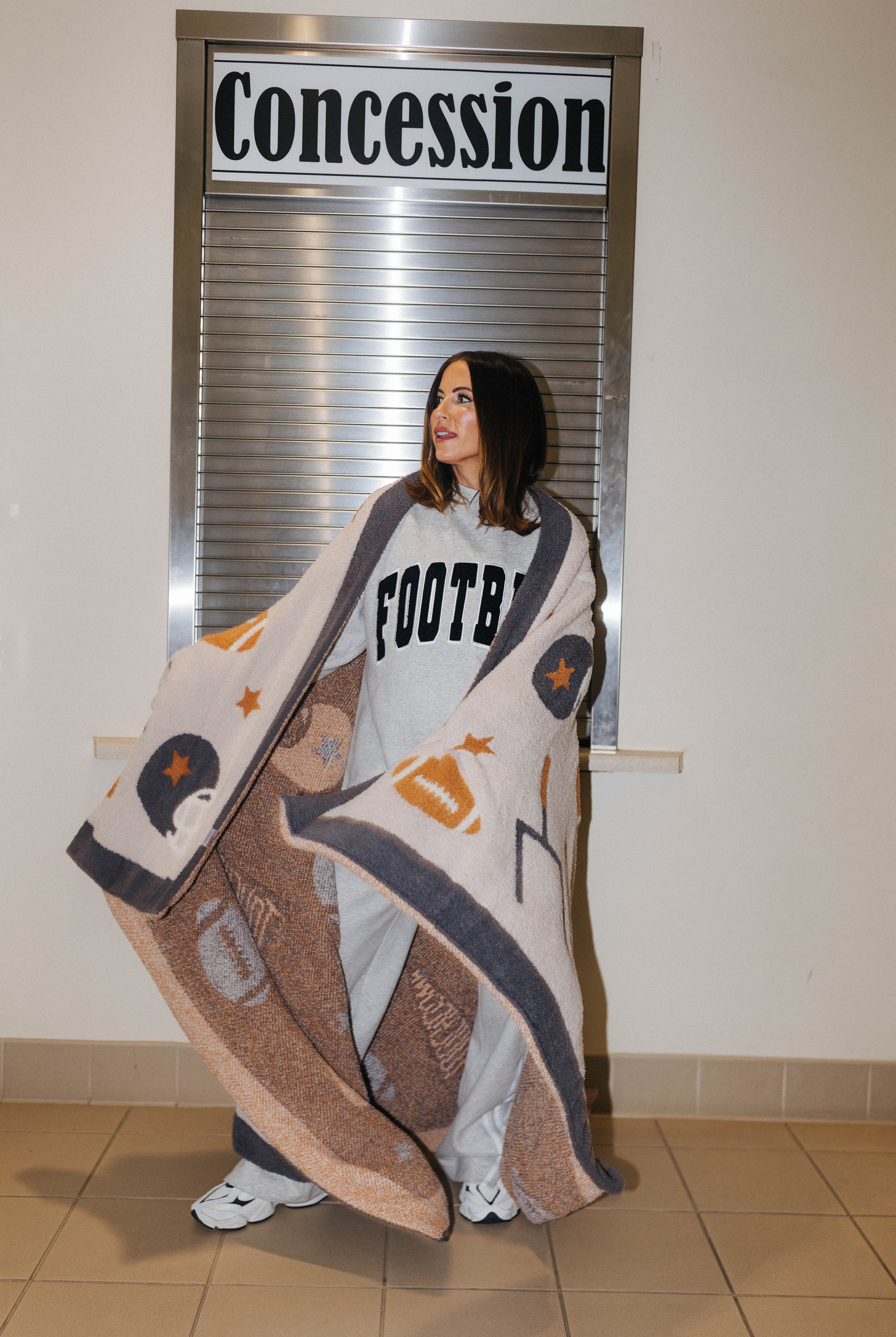 A woman stands indoors in front of a window with a 'Concession' sign, wrapped in a large beige and brown football-themed blanket with grey and orange accents. She is wearing a light gray sweatshirt with 'FOOTBALL' printed on it.