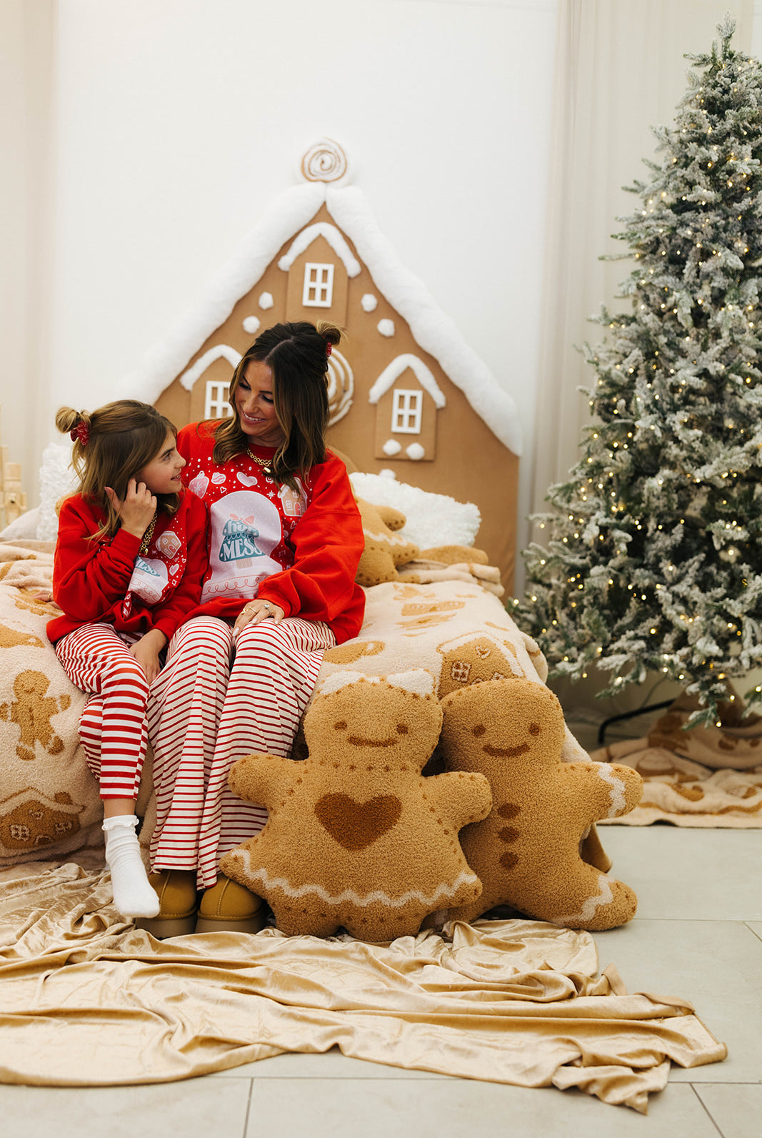 Two people in pajamas sitting on a bed with gingerbread-themed decor and a Christmas tree in the background.