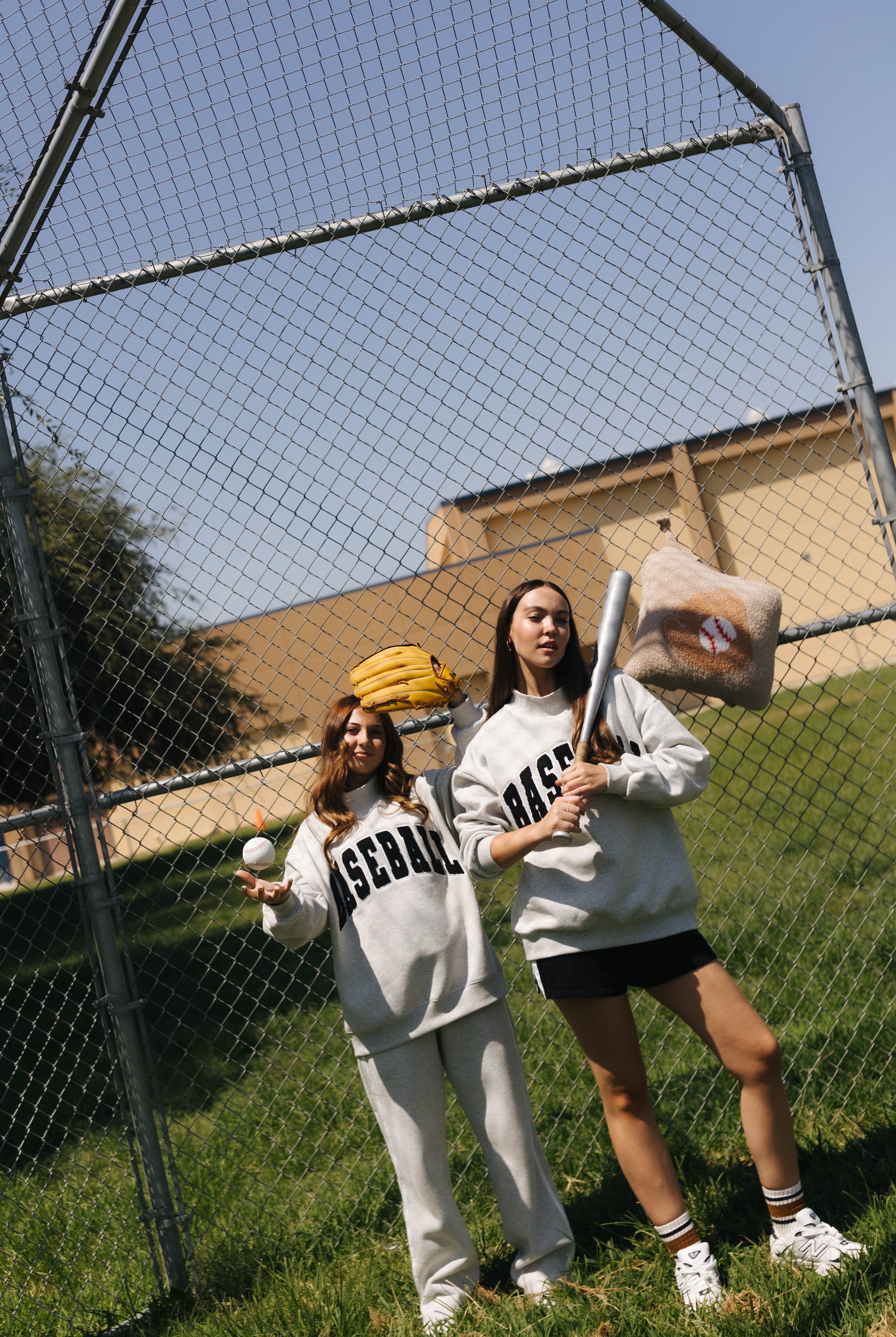 Two women posing in a baseball field wearing matching light gray sweatpants and sweatshirts with 'BASEBALL' printed in black. One holds a baseball bat and the other holds a baseball, with a chain-link fence and grassy field in the background.