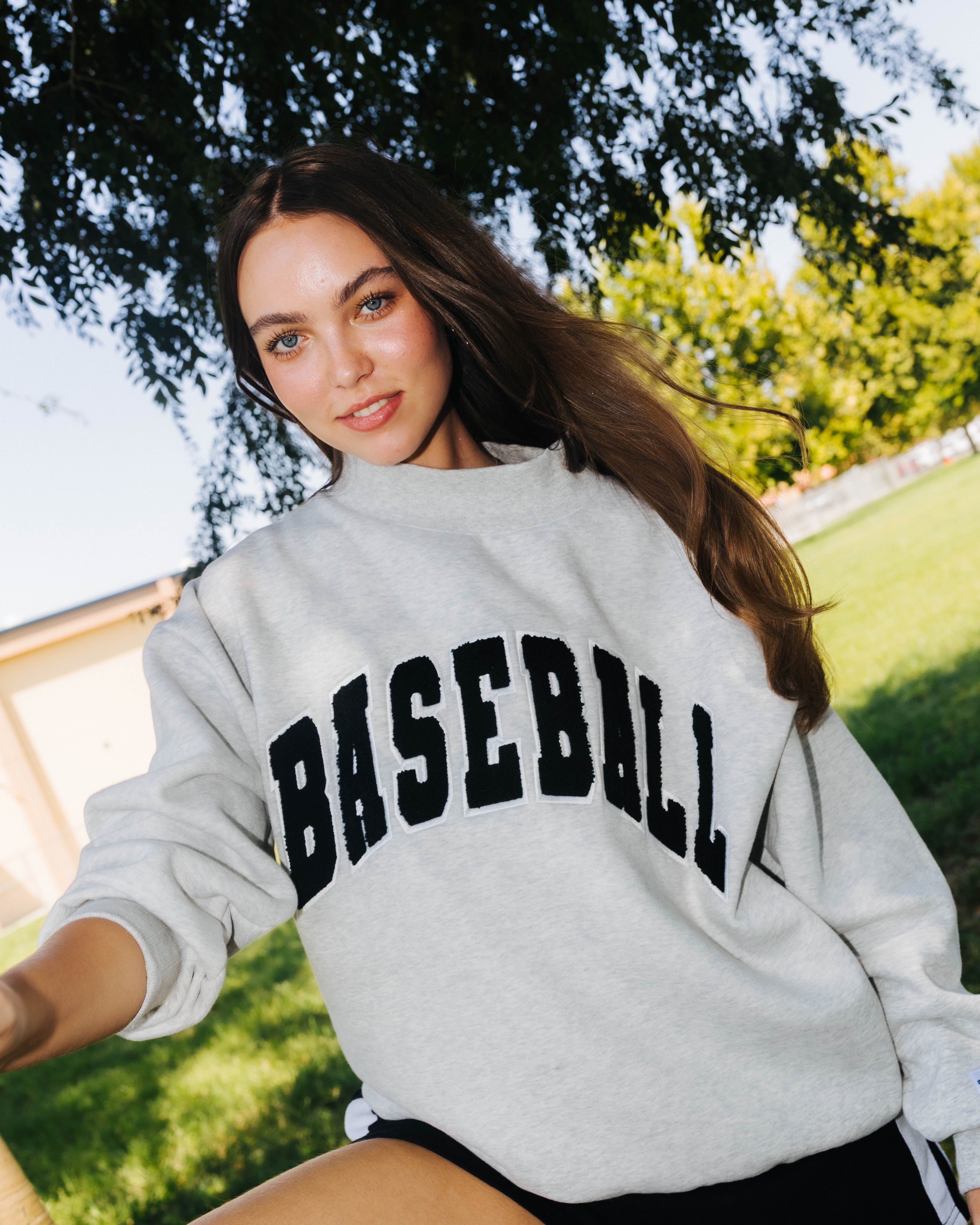 A woman outdoors wearing a light gray sweatshirt with 'BASEBALL' printed across the front in black arched lettering, paired with black shorts.