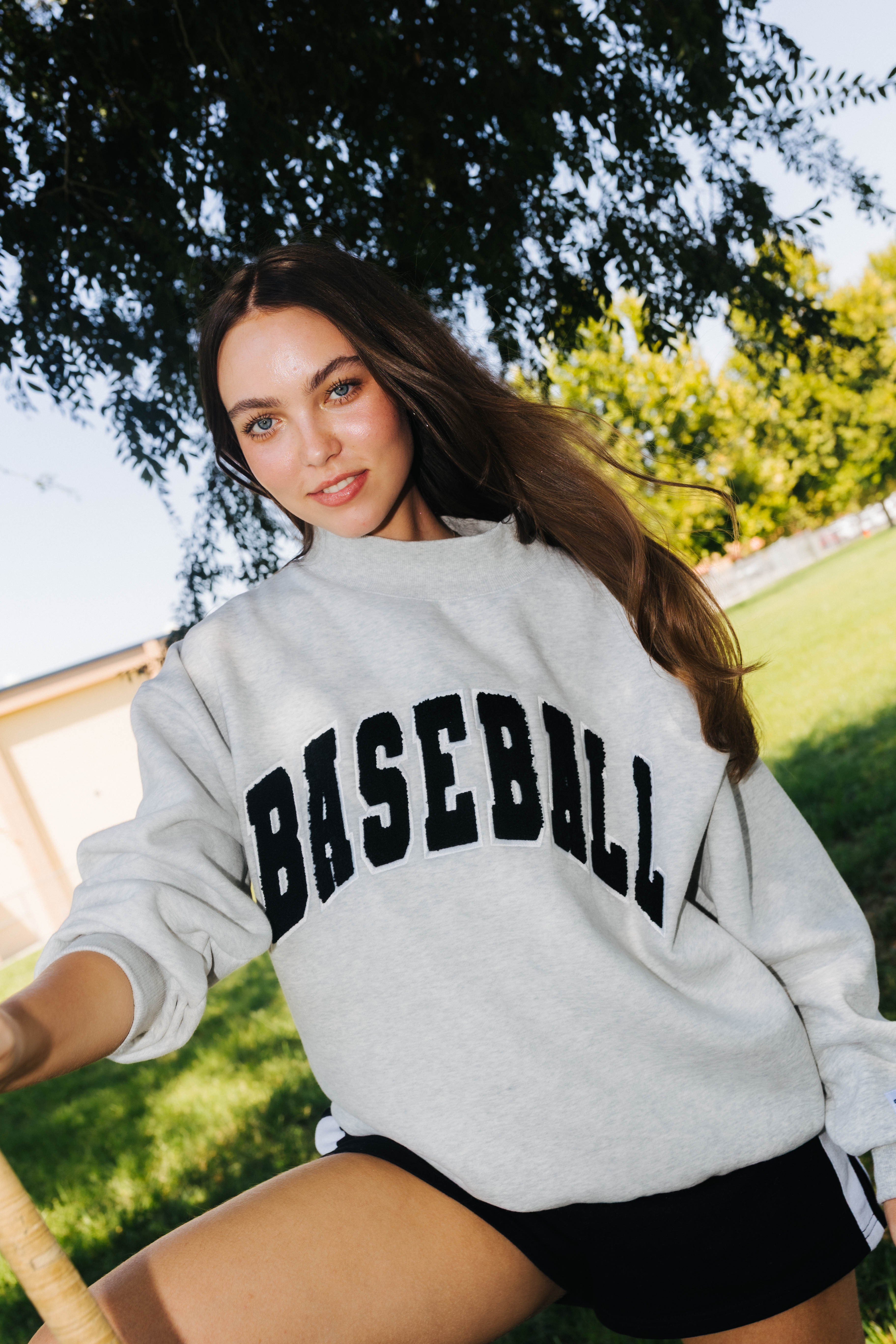 A woman outdoors wearing a light gray sweatshirt with 'BASEBALL' printed across the front in black arched lettering, paired with black shorts.