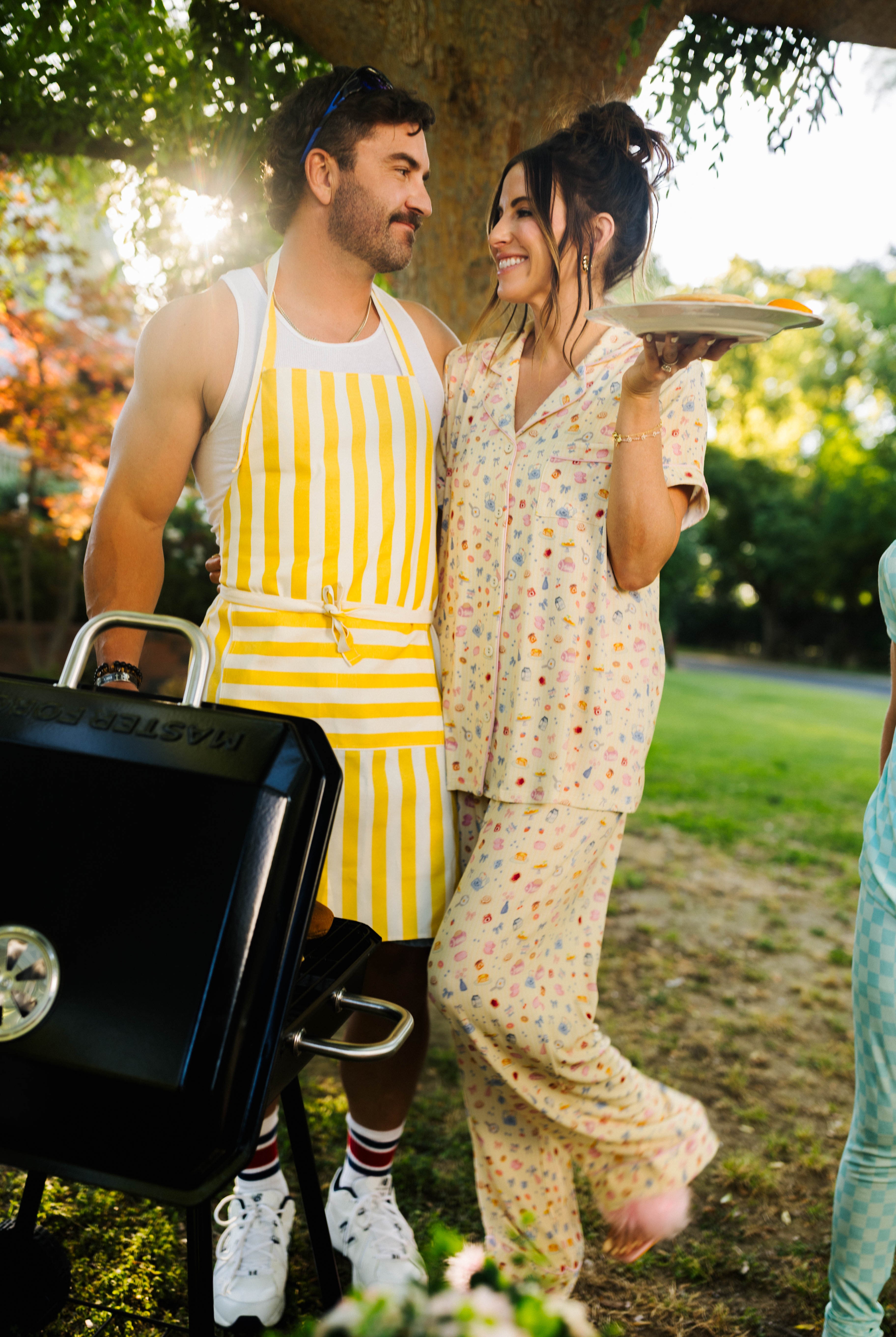 Man and woman standing outdoors with a grill and food, surrounded by greenery.