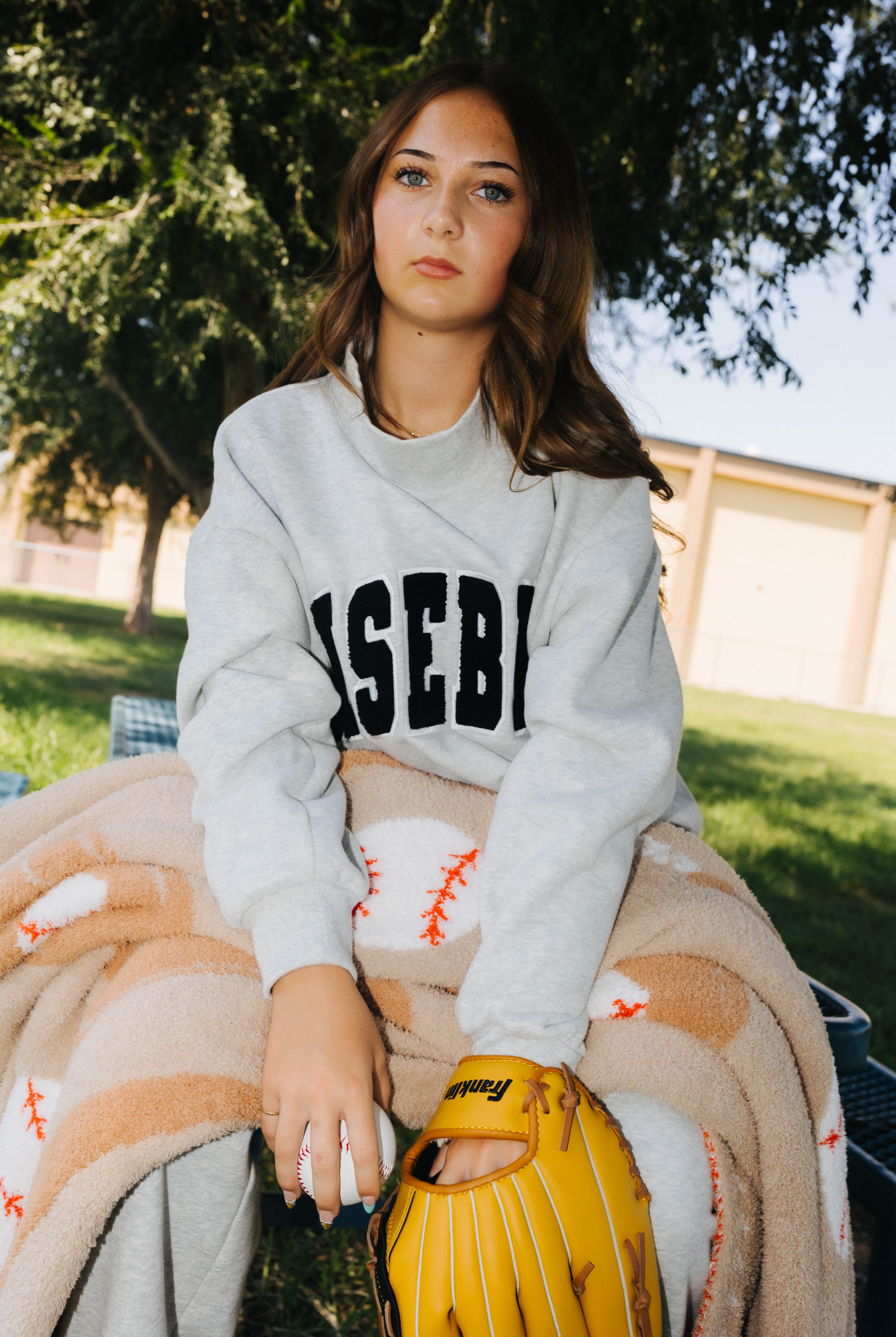A woman sitting outdoors on a blanket, wearing a light gray sweatshirt with 'BASEBALL' printed in black across the chest, holding a yellow baseball glove.