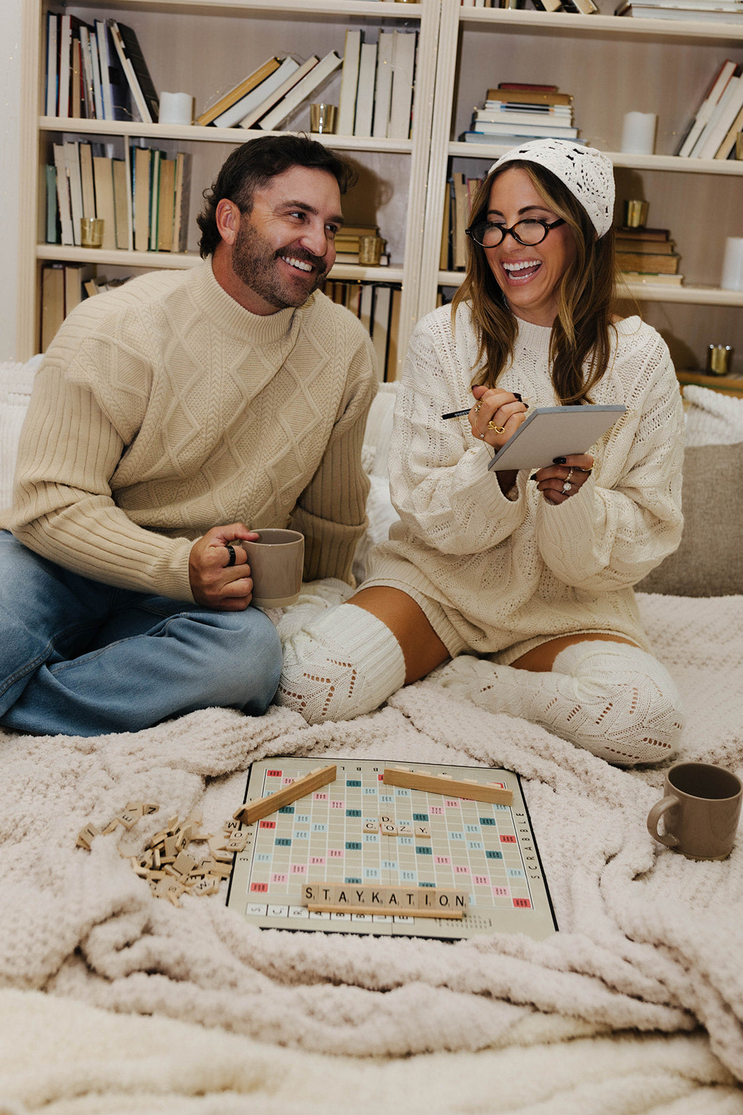 Man and woman sitting on a bed with a Scrabble board and tablet, surrounded by books.