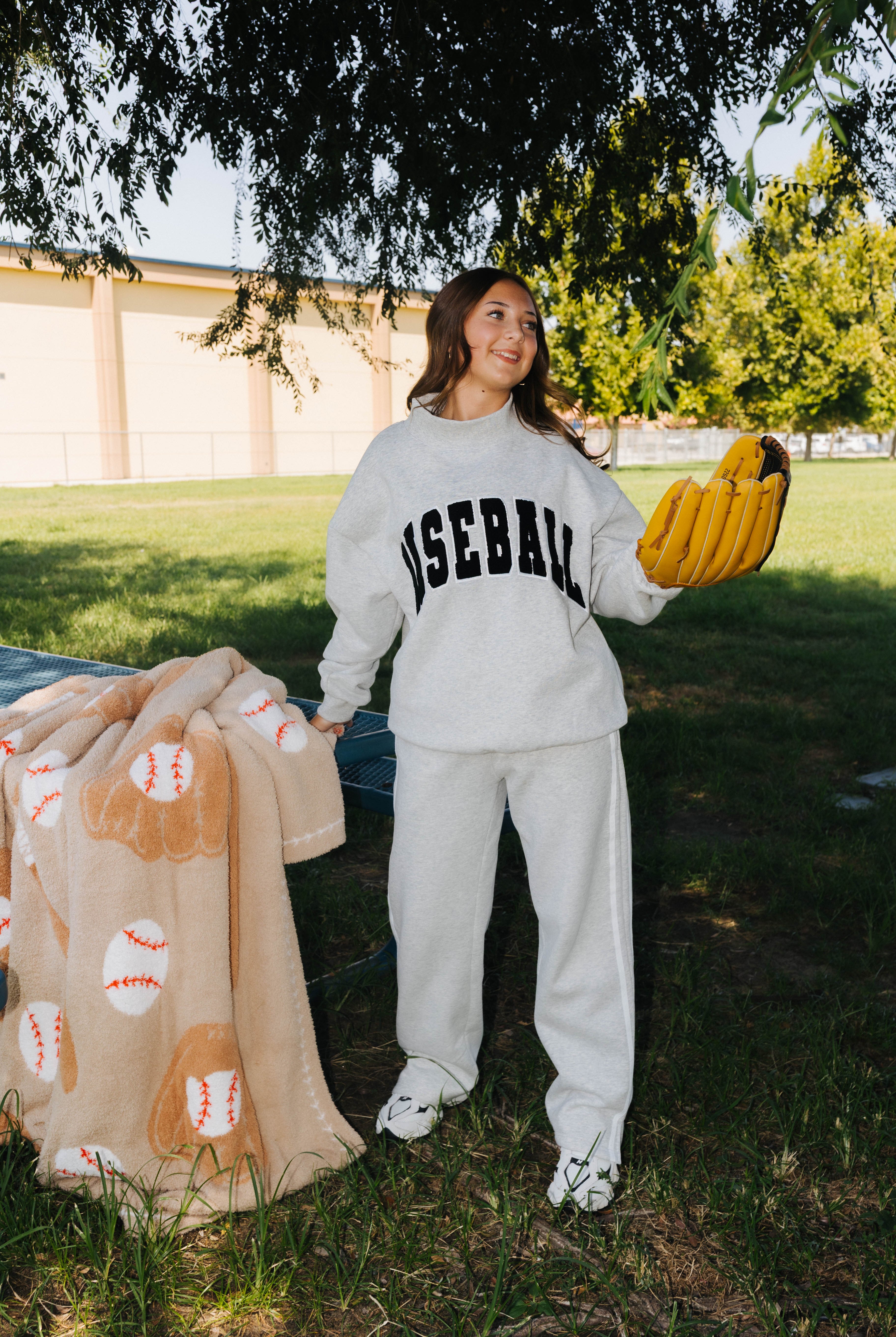 A woman stands outdoors on grass under a tree wearing a light gray baseball-themed sweatshirt and sweatpants set, holding a tan baseball-patterned blanket and a yellow glove.