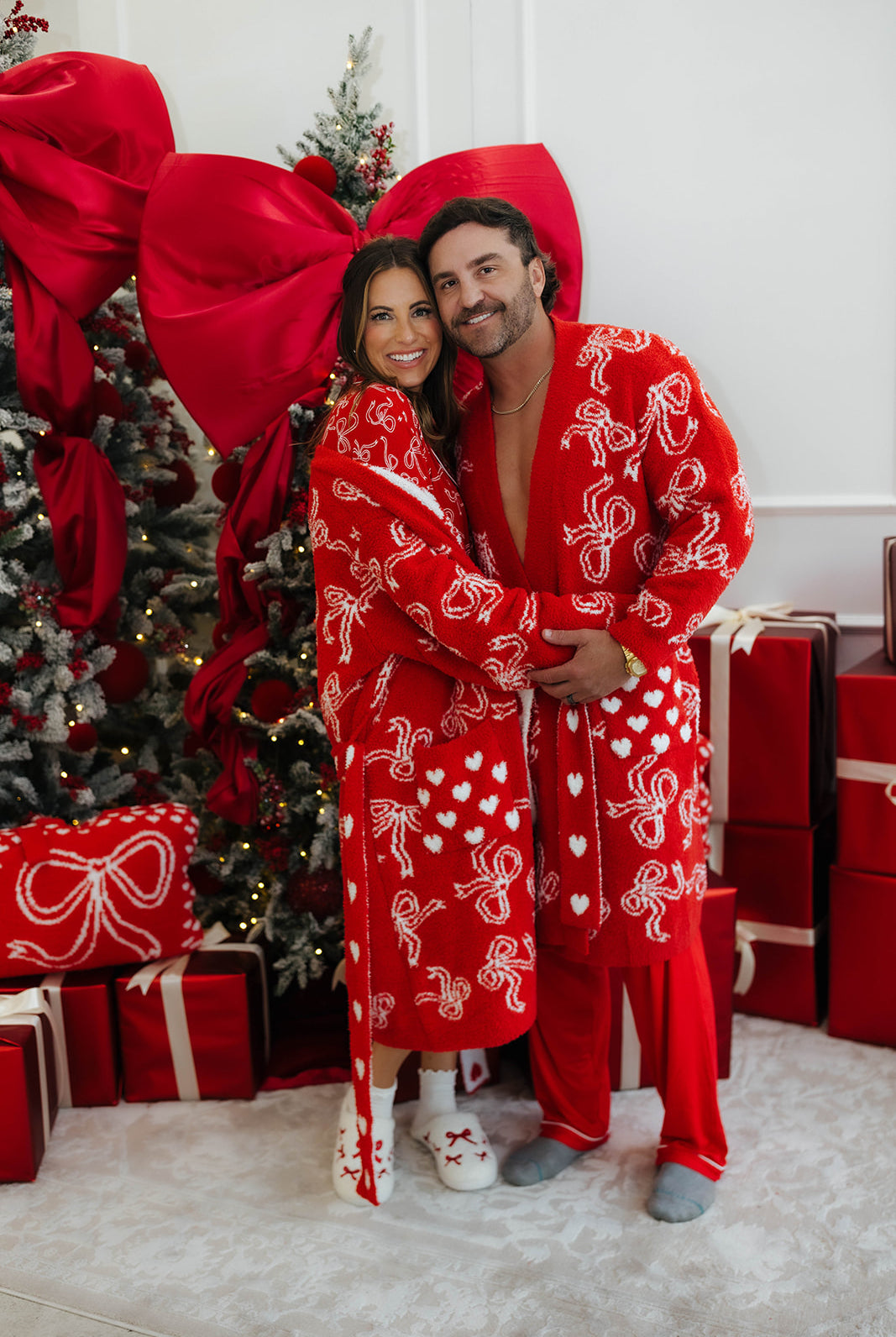 Couple in matching red Christmas robes standing in front of a decorated Christmas tree and presents.