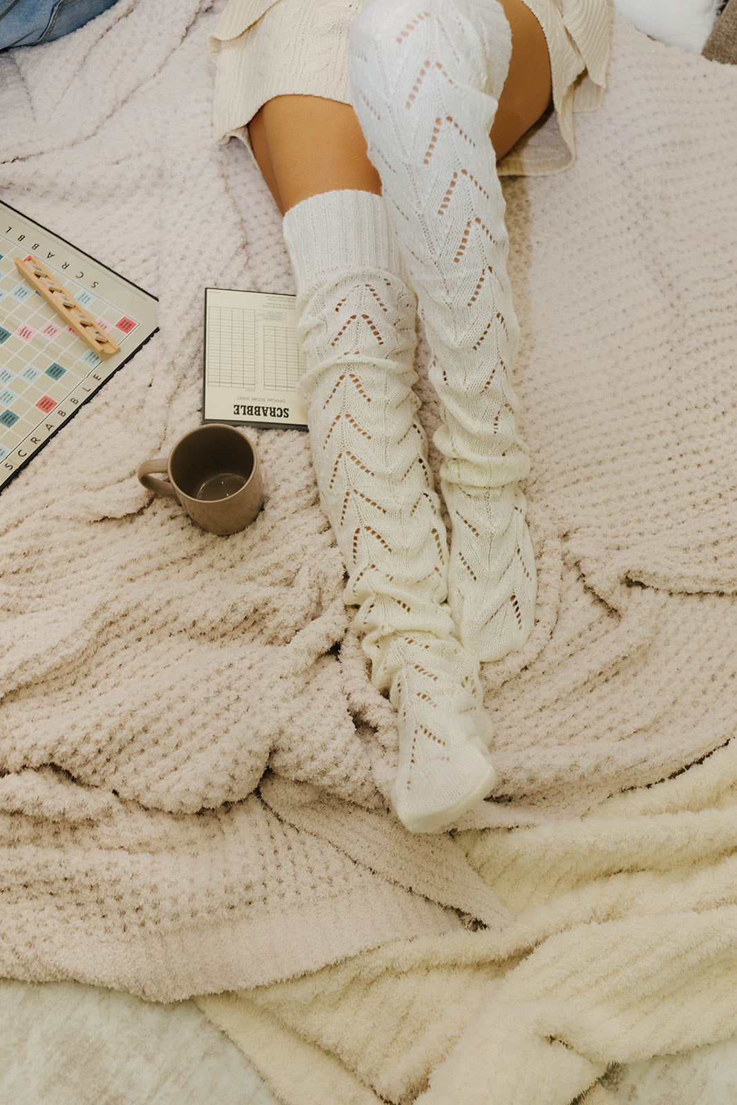 Person wearing white knitted knee-high socks on a cozy blanket with a mug and book.