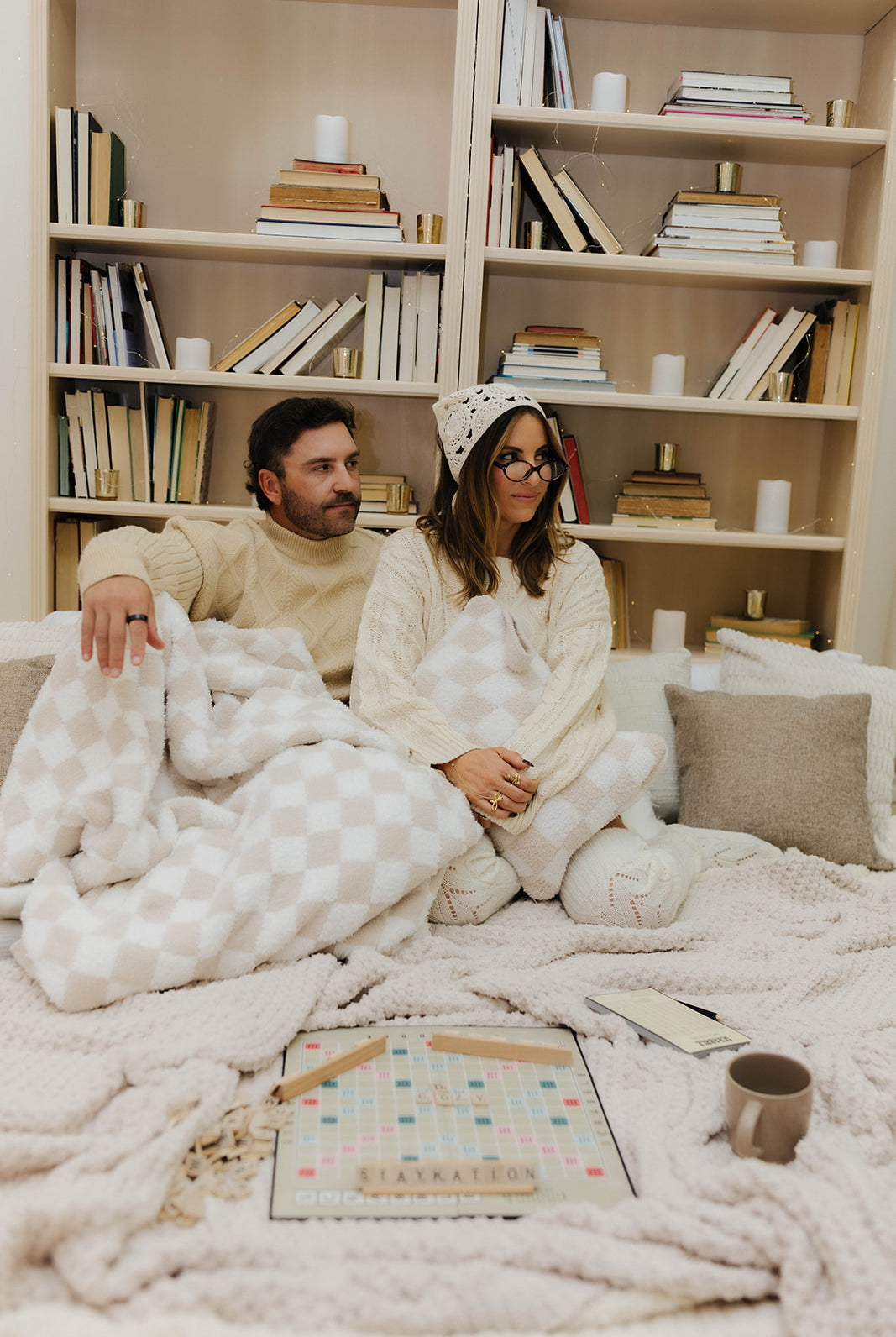 Two people sitting on a couch under a blanket with a bookshelf in the background