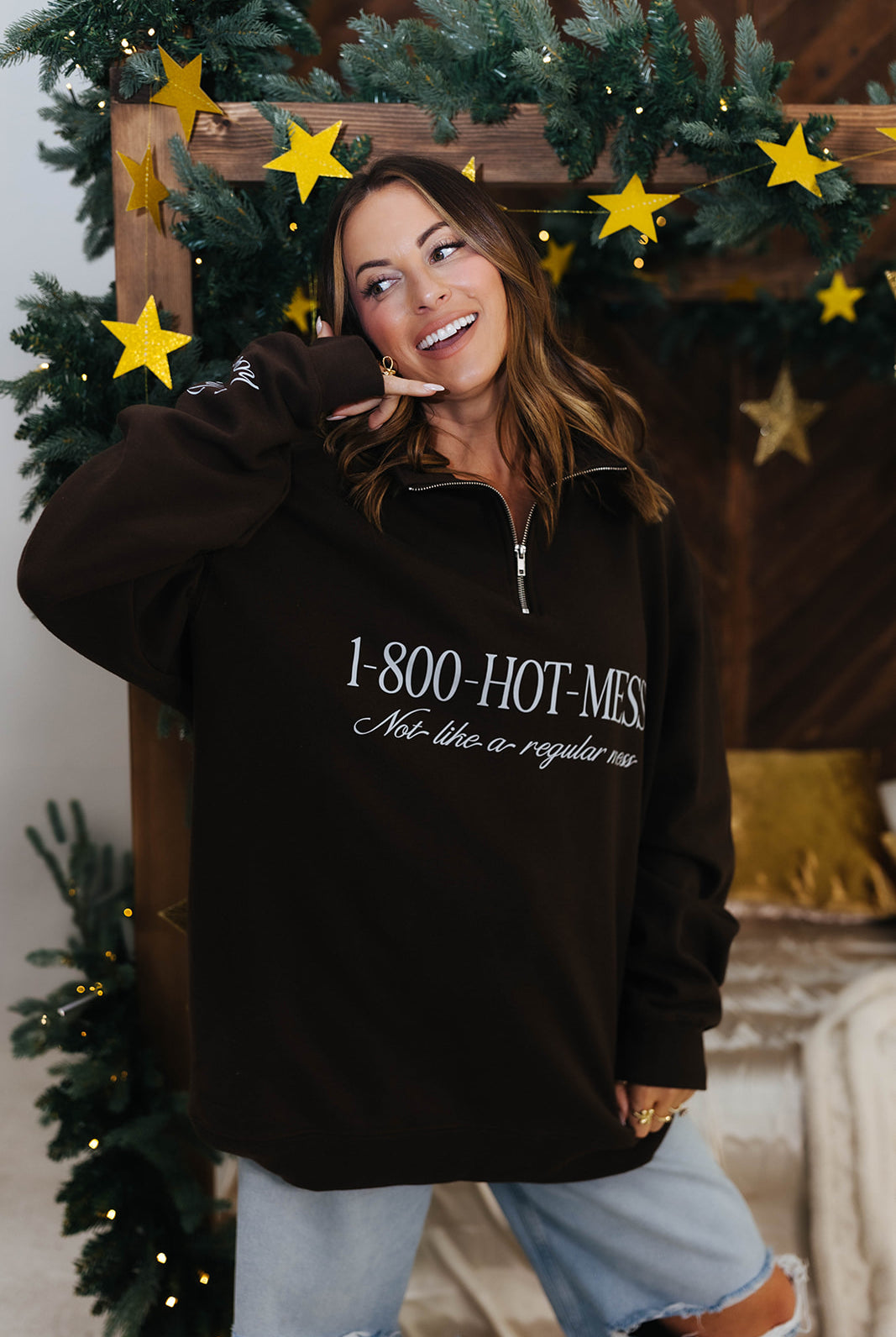 Person wearing a brown sweatshirt with text, standing in front of a decorated Christmas tree.