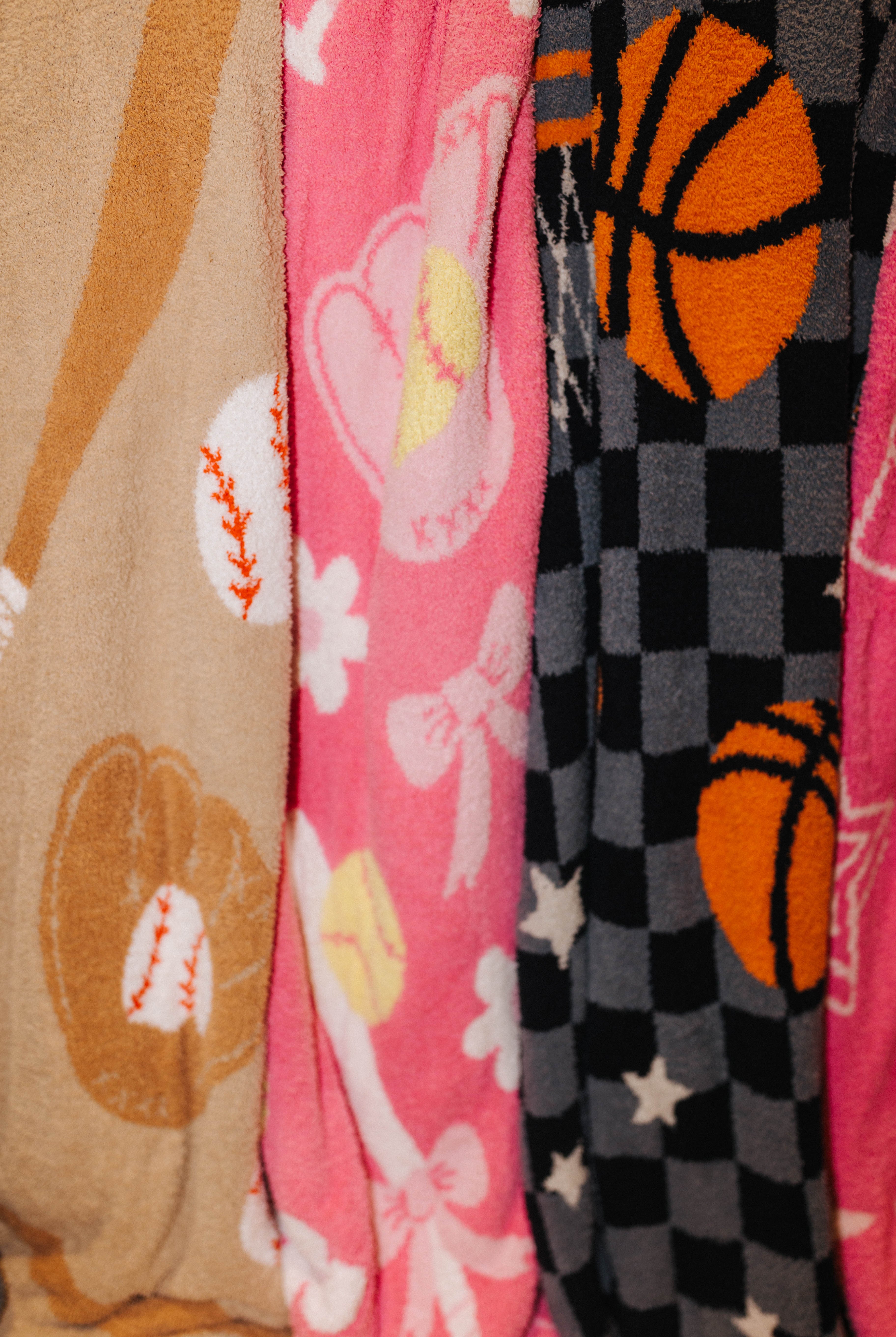 Close-up of three patterned fabric items hanging side by side, including a tan blanket with baseball motifs, a pin blanket with bows and softballs, and a dark gray/black checkered fabric with orange basketballs and white stars.