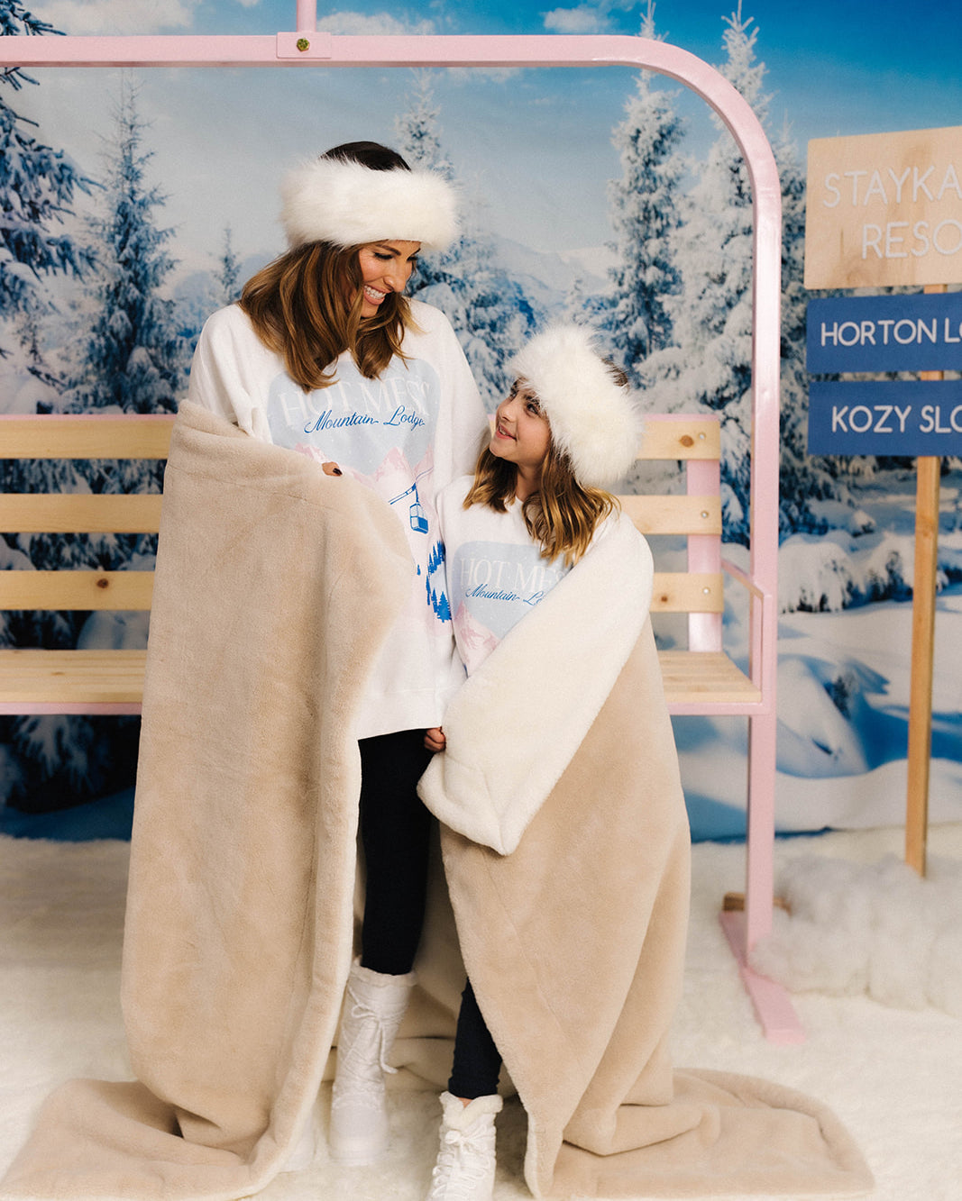 Two people wrapped in a blanket in a snowy landscape with a pink structure and signs in the background.