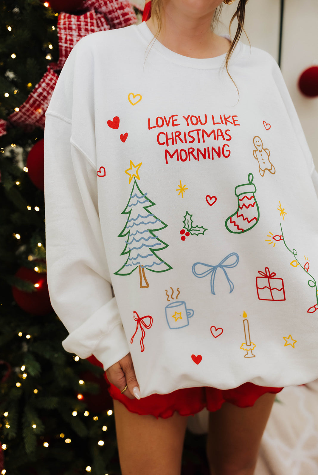 Person wearing a white sweatshirt with Christmas-themed designs and text in front of a decorated Christmas tree.