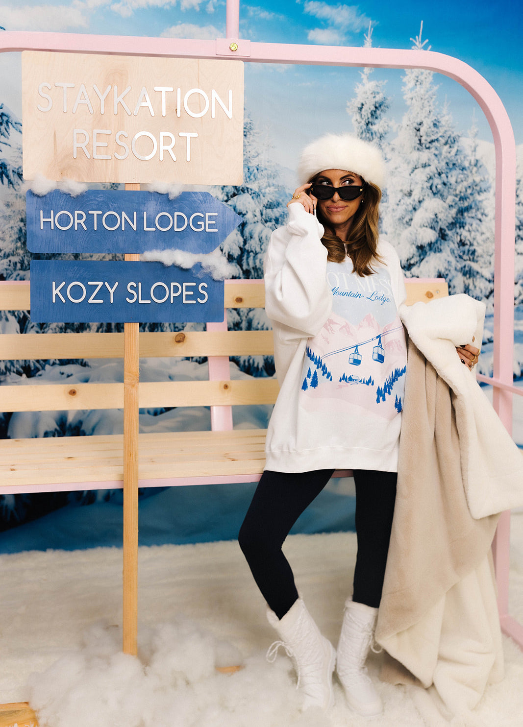 Person in winter clothing standing in front of a ski resort sign with snowy trees in the background