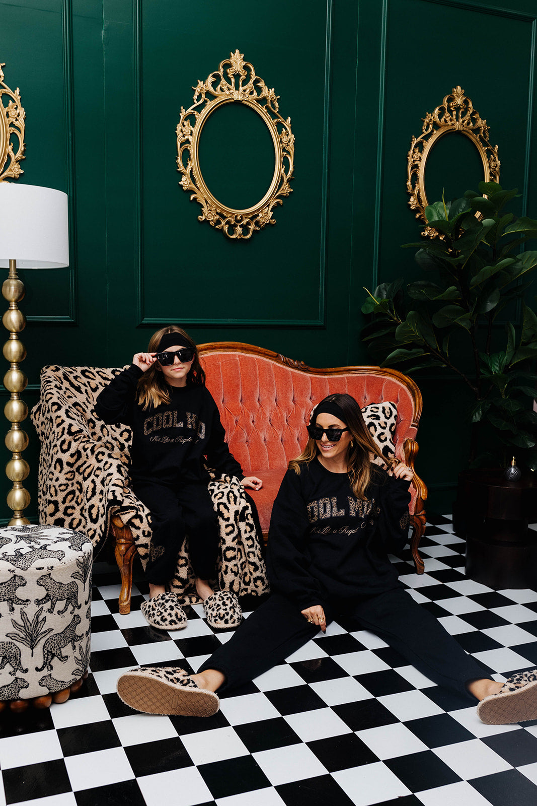Two women sitting on a checkered floor in a room with ornate gold mirrors and a red couch.