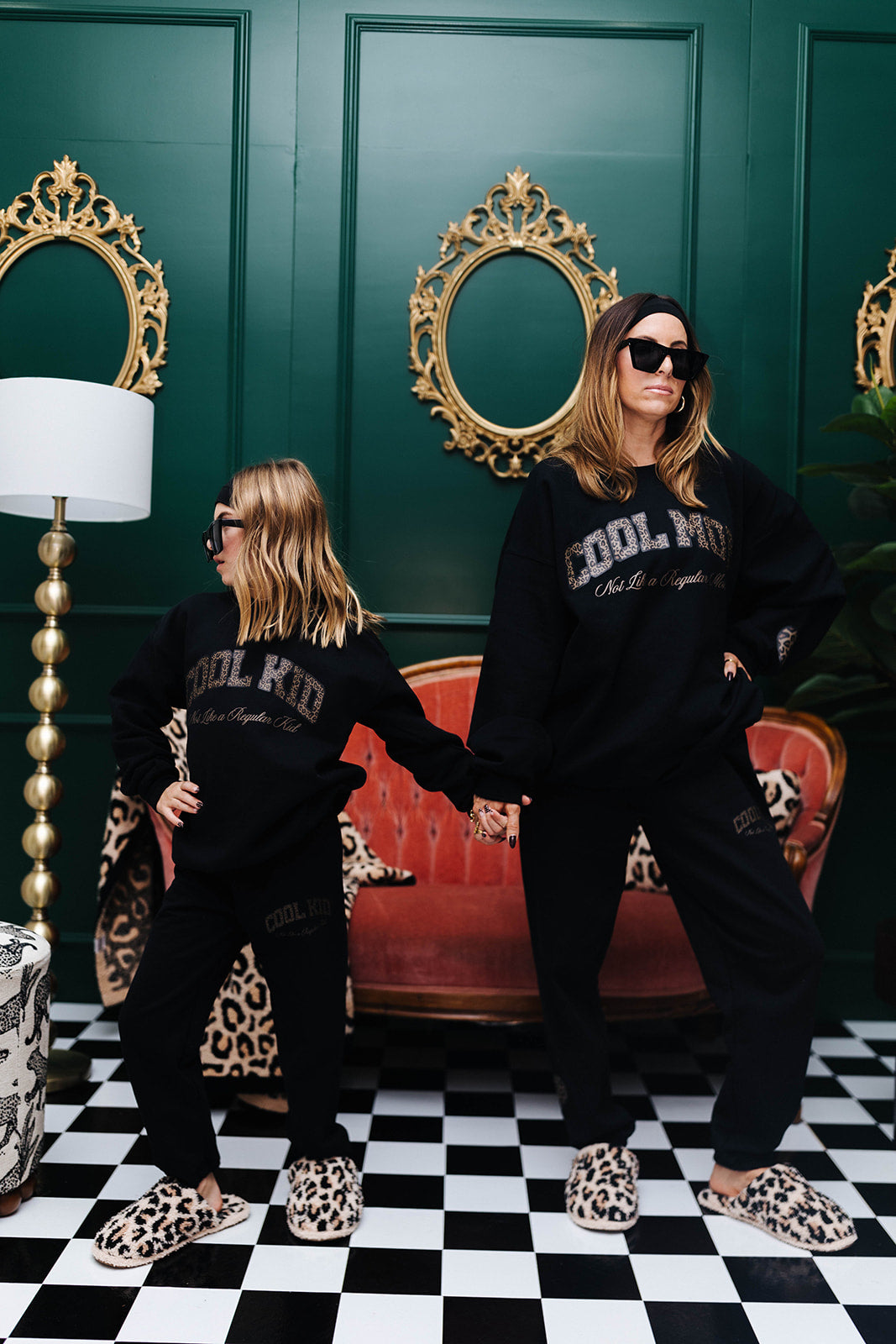 Two people wearing matching black sweatshirts with 'COOL KID' text in a stylish room with green walls and decorative mirrors.