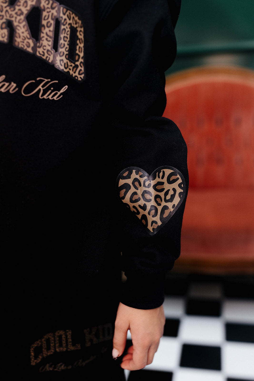 Black hoodie with leopard print heart design on sleeve, worn by a person, with a checkered floor and red chair in the background.