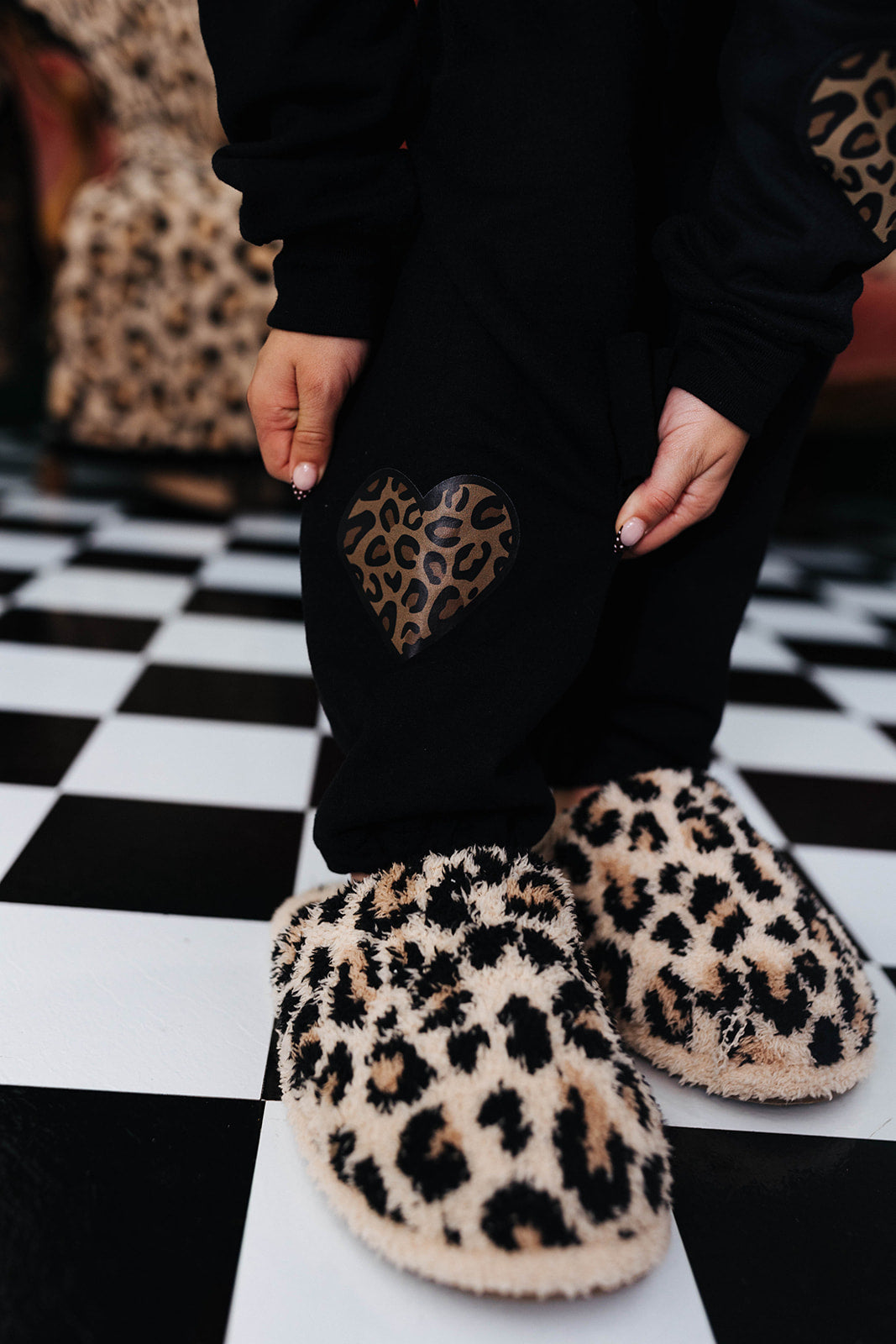 Person wearing black pants with a heart design and leopard print slippers on a checkered floor.