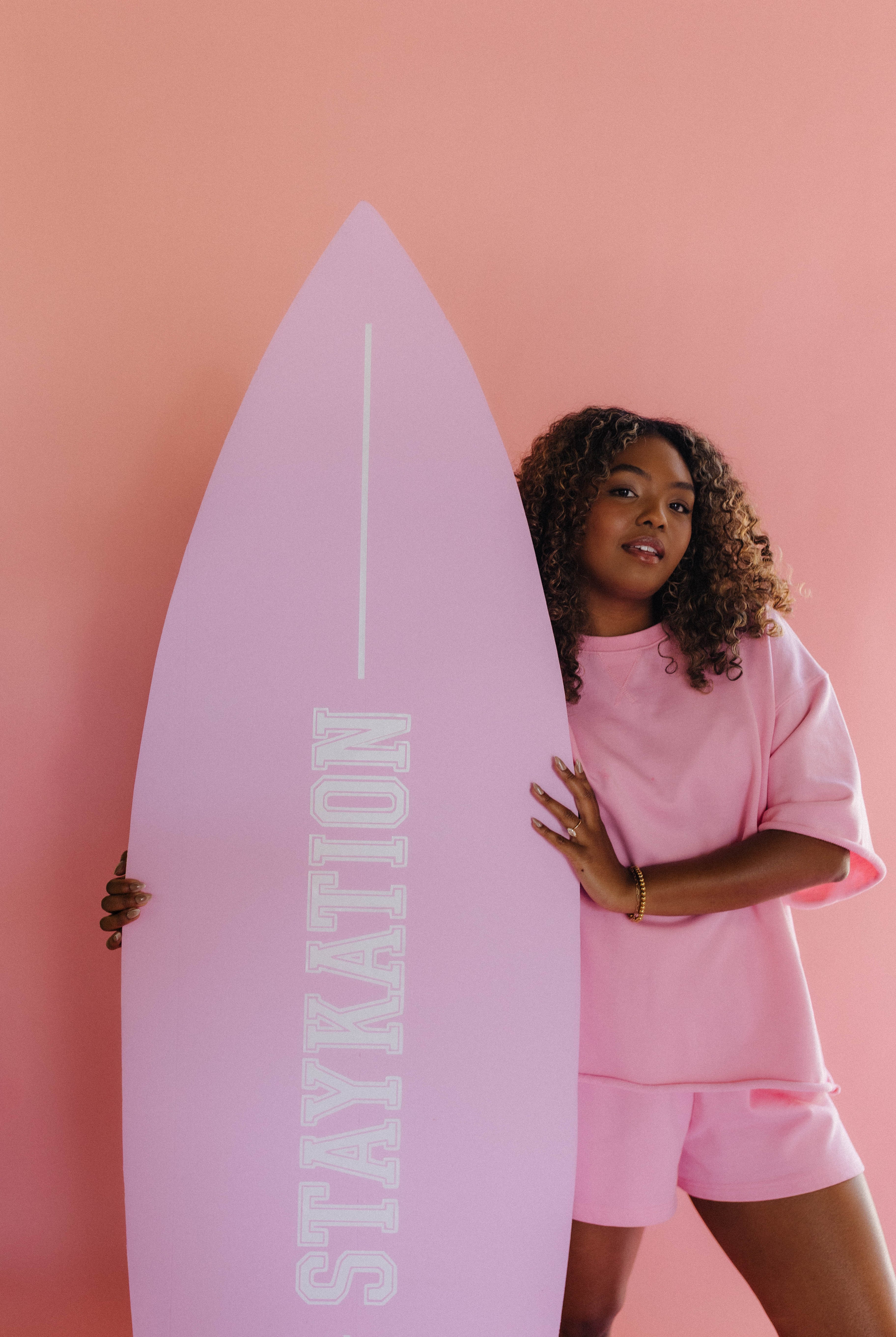 Person holding a pink surfboard with 'STAYKATION' text against a pink background