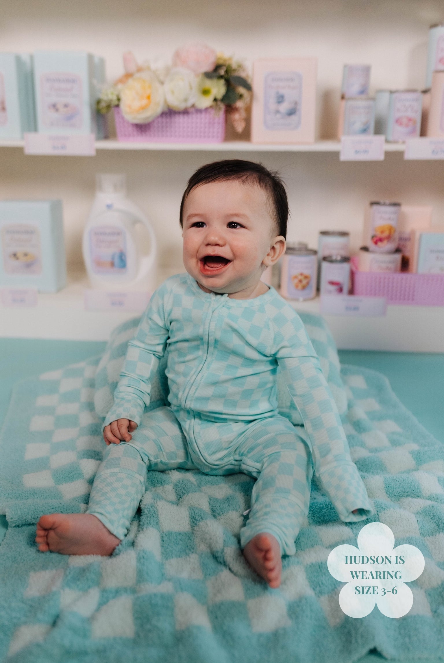 Baby in a teal outfit sitting on a checkered blanket with shelves of products in the background