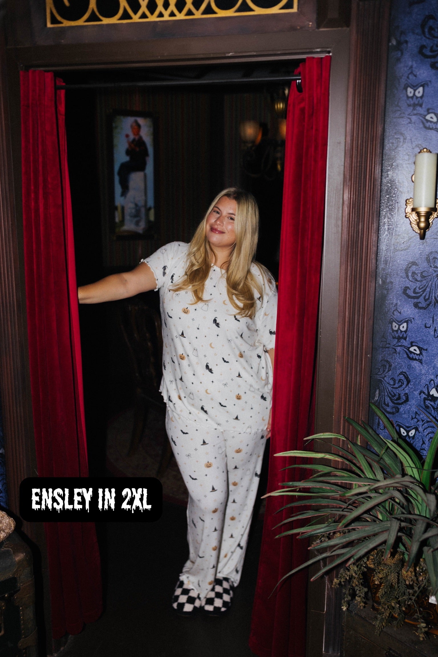 A woman wearing a white short-sleeve pajama set with a black and gold bat pattern stands in a doorway framed by red curtains. The text overlay at the bottom left reads 'ENSLEY IN 2XL'.
