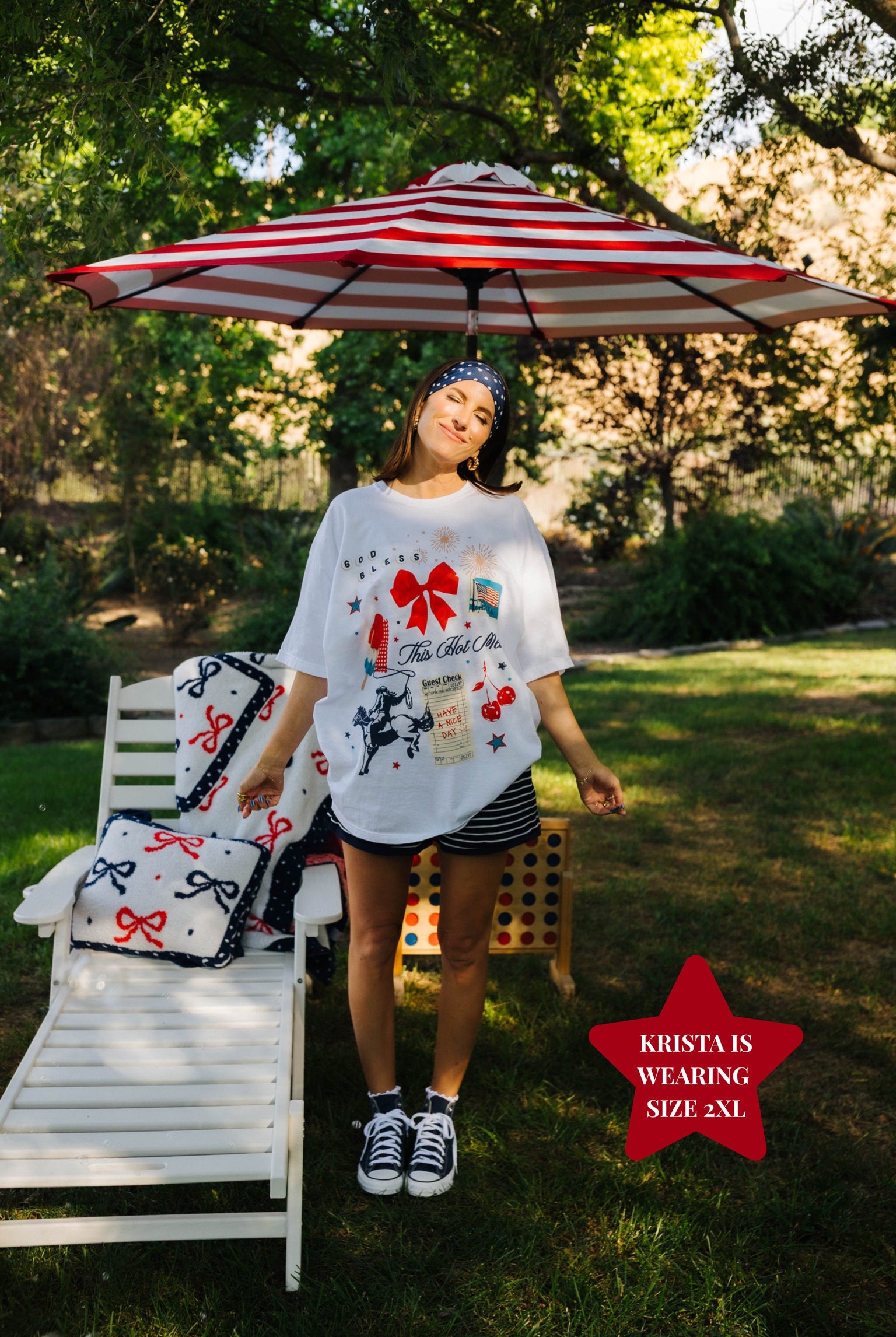 Woman standing under a red and white striped umbrella with a garden chair and decorative pillows in the background.