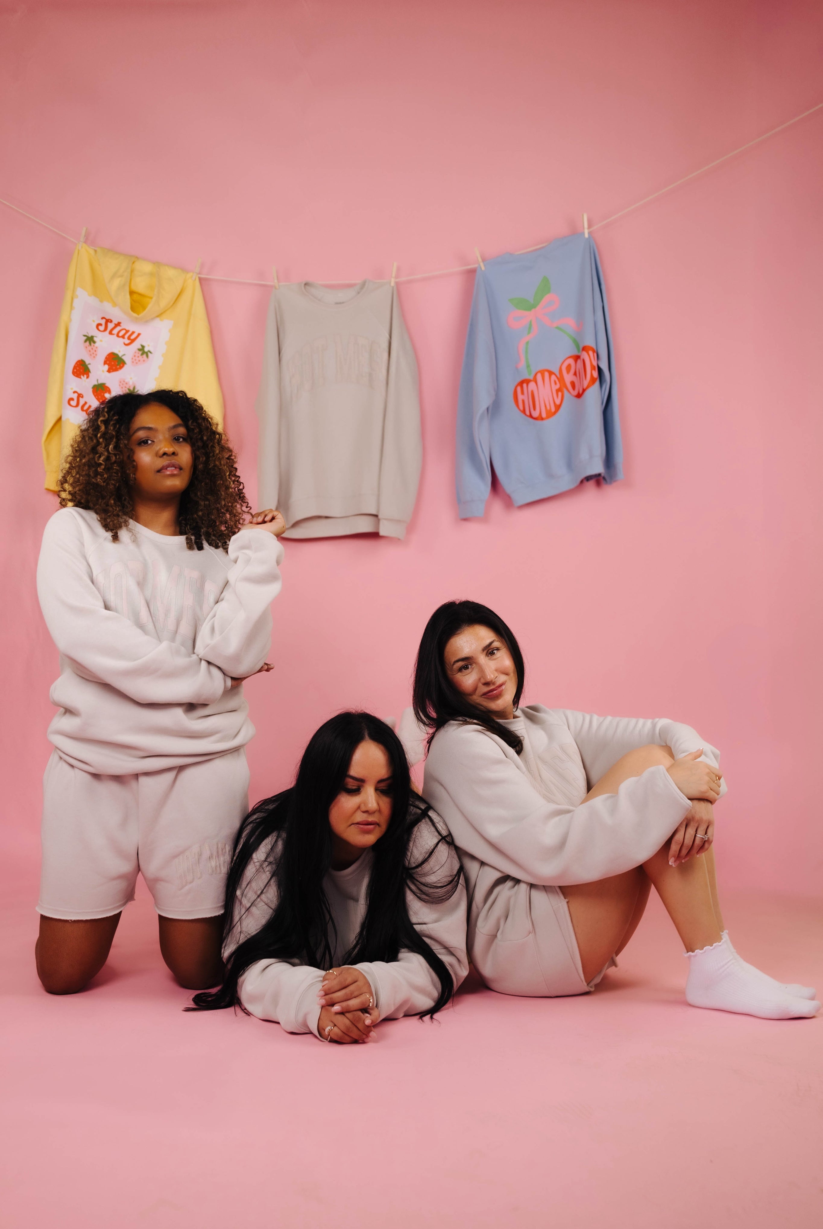 Three women posing on a pink background with colorful sweatshirts hanging on a line.