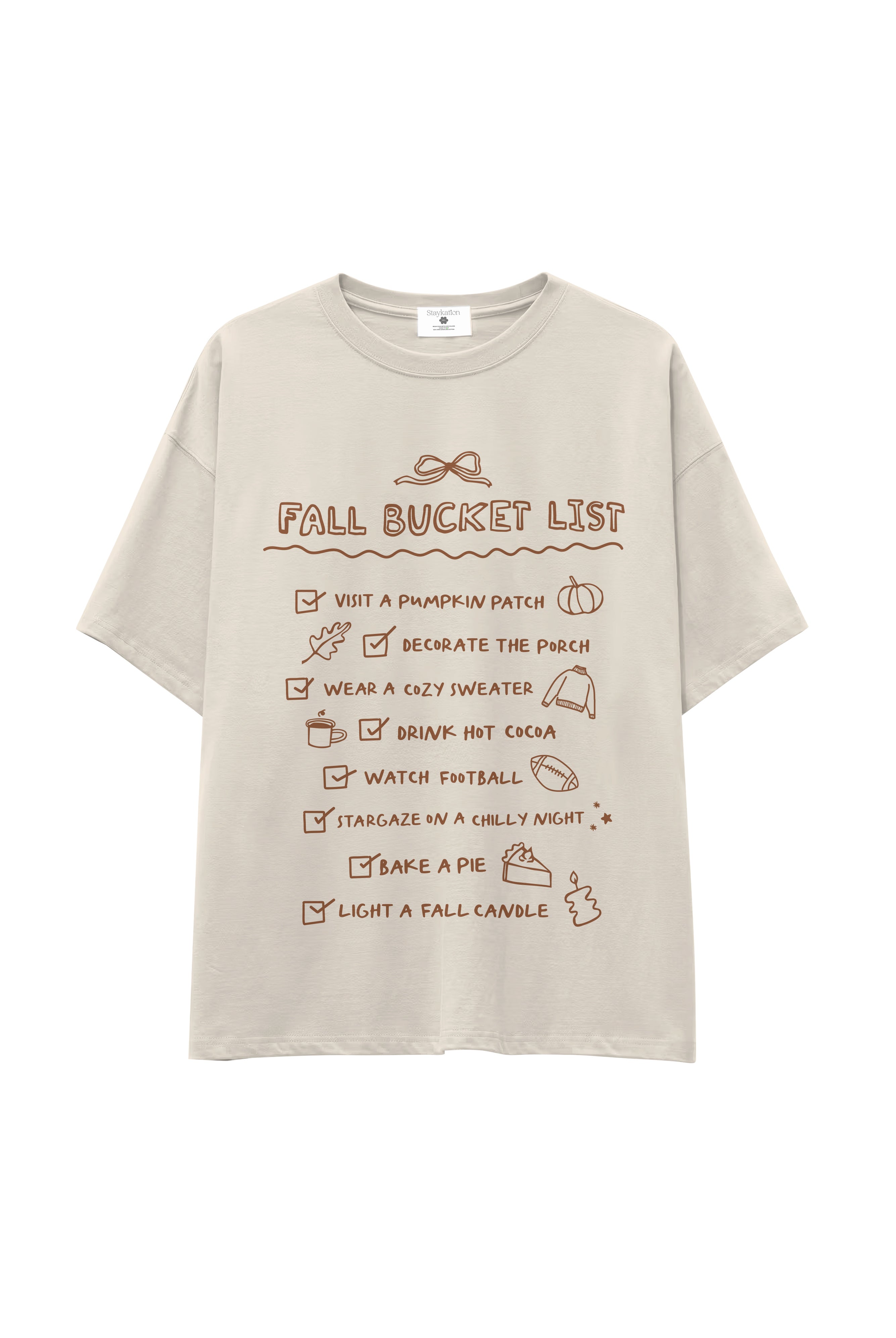 A light beige oversized t-shirt with a brown graphic print titled 'FALL BUCKET LIST' featuring various autumn-themed checklist items, shown against a plain white background.