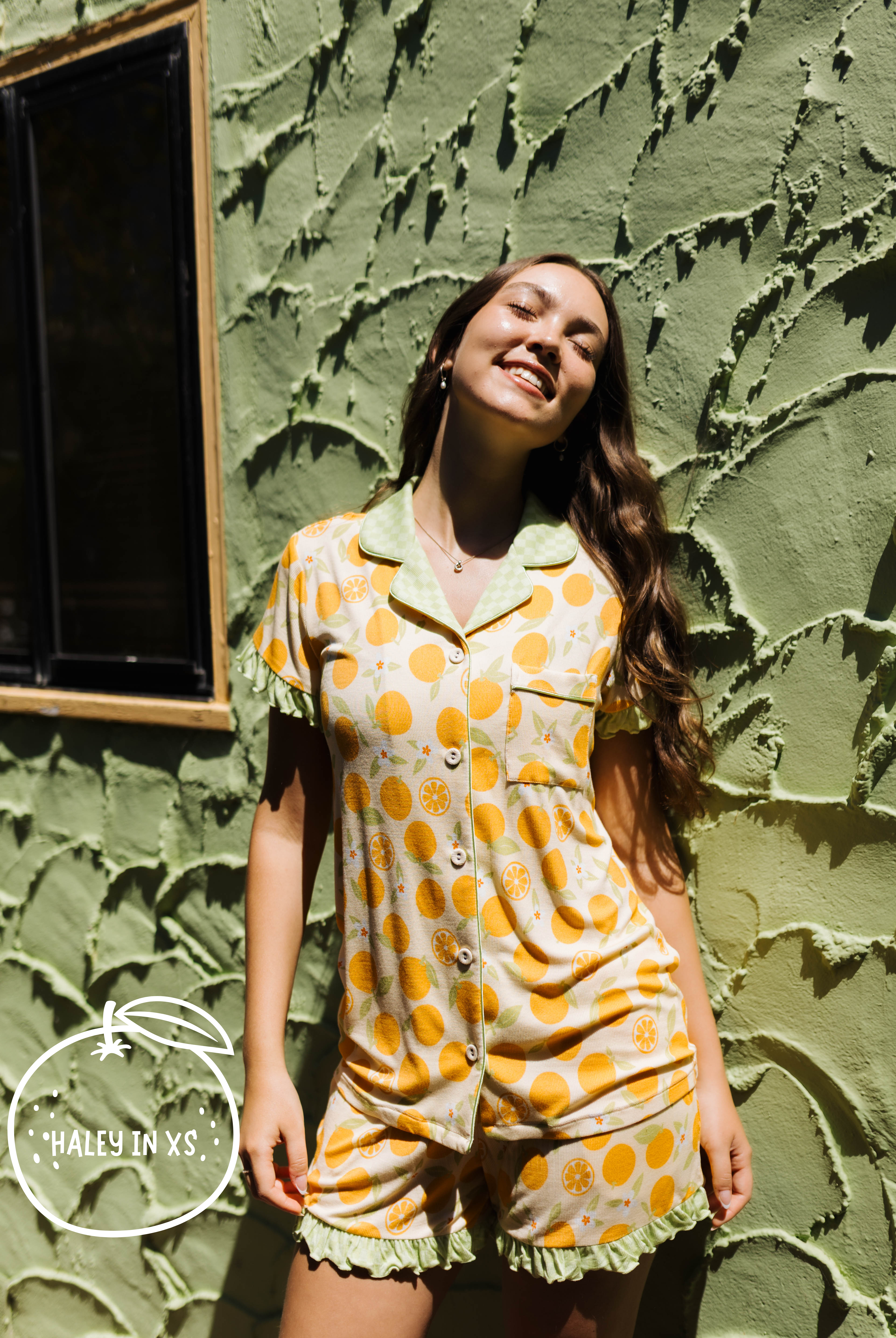 A woman wearing a short-sleeve button-front pj matching ruffled shorts, featuring orange citrus fruit print. She is posing outdoors against a textured olive green stucco wall.
