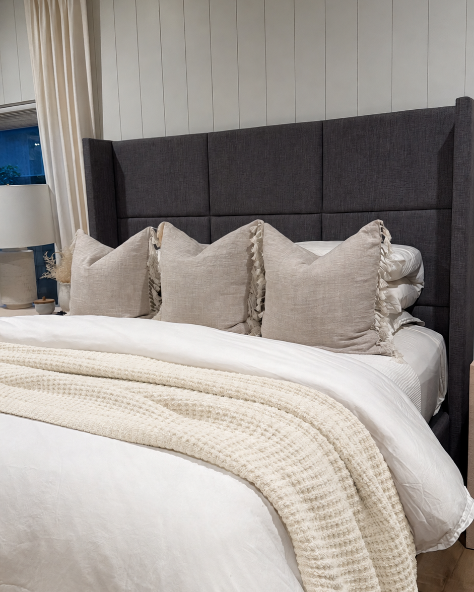 A styled bedroom scene showing a bed with a tall dark gray upholstered headboard, white bedding, beige pillows, and a cream textured throw blanket. The wall behind the bed has vertical white paneling.
