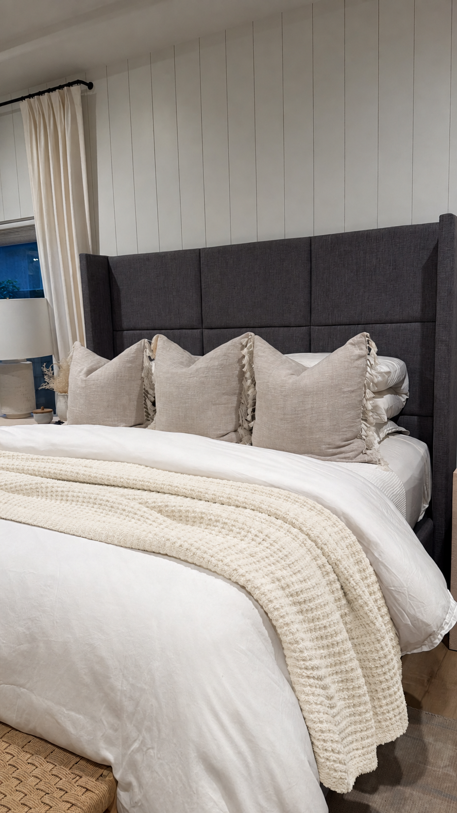 A styled bedroom scene showing a bed with a tall dark gray upholstered headboard, white bedding, beige pillows, and a cream textured throw blanket. The wall behind the bed has vertical white paneling.