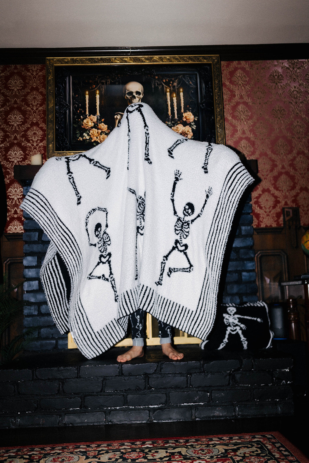 A person standing barefoot in a dimly lit room, wrapped in a large white blanket printed with black skeleton figures and striped borders. The room has dark brick hearth, red patterned wallpaper, and a framed artwork above.