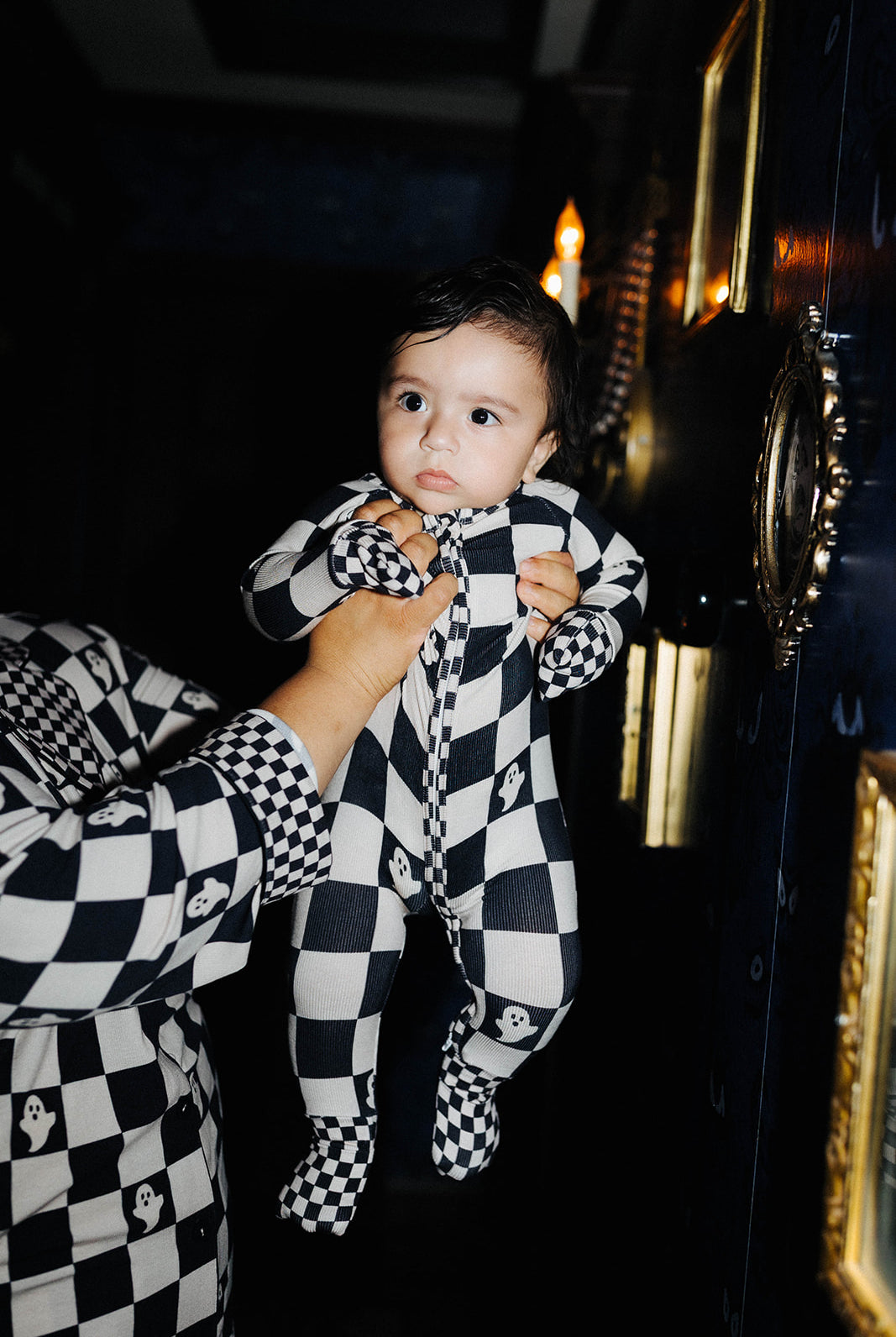 A baby wearing a black and sand checkered footed onesie is held up by an adult wearing a matching checkered shirt in a dark interior setting.