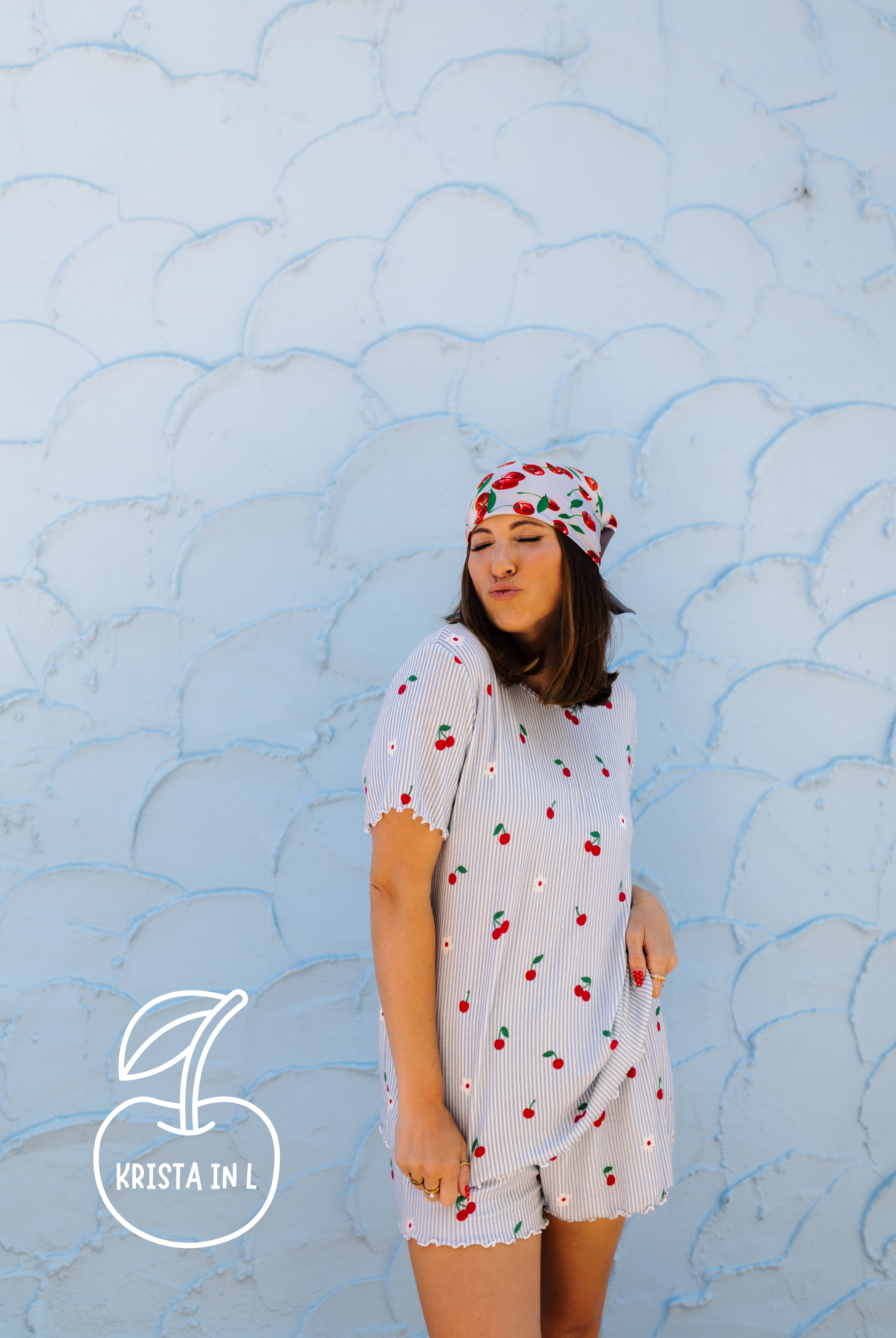 A woman wearing a white cherry-patterned pj and matching headscarf stands against a light blue textured wall. The brand logo 'Krista in L' with an cherry icon is visible in the bottom-left corner.
