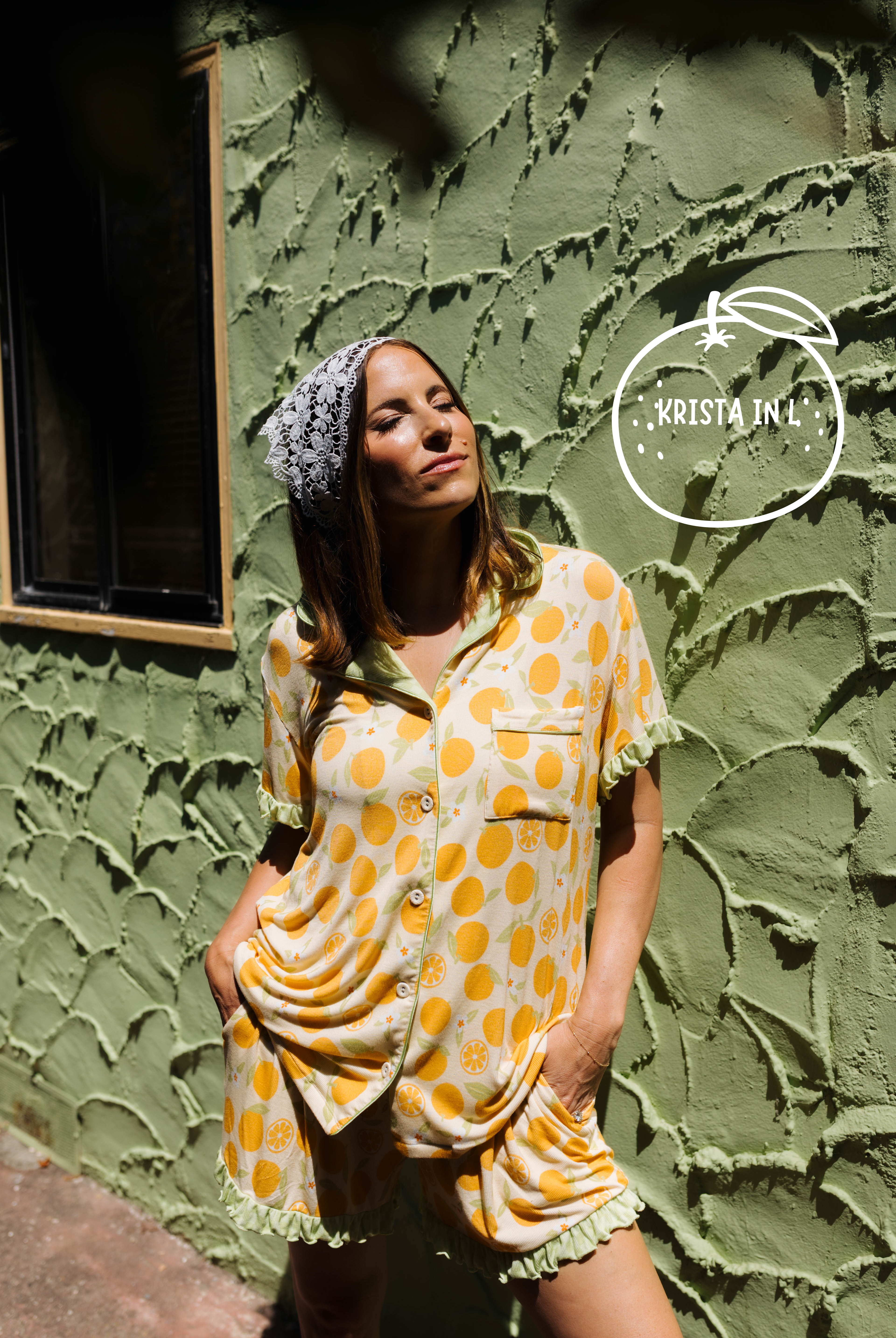 A woman wearing an orange pajama set with a matching stands against a textured olive green stucco wall. A white 'KRISTA IN A' logo watermark is visible in the upper right.