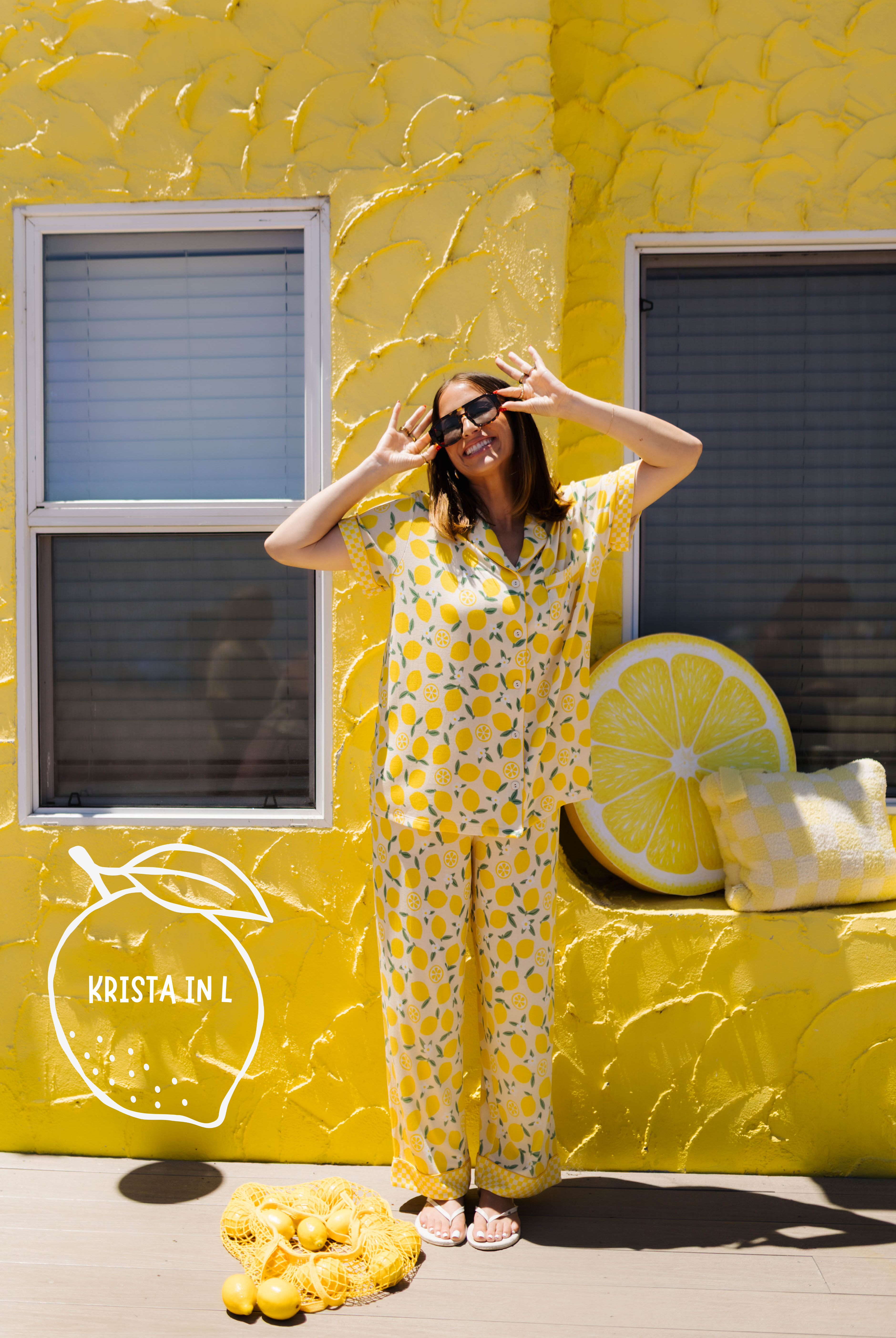 A woman wearing a yellow lemon-patterned pajama set stands in front of a bright yellow textured wall. The scene includes a large lemon slice prop on the wall, a pile of lemons on the ground, and a logo overlay reading 'Kristin L'.