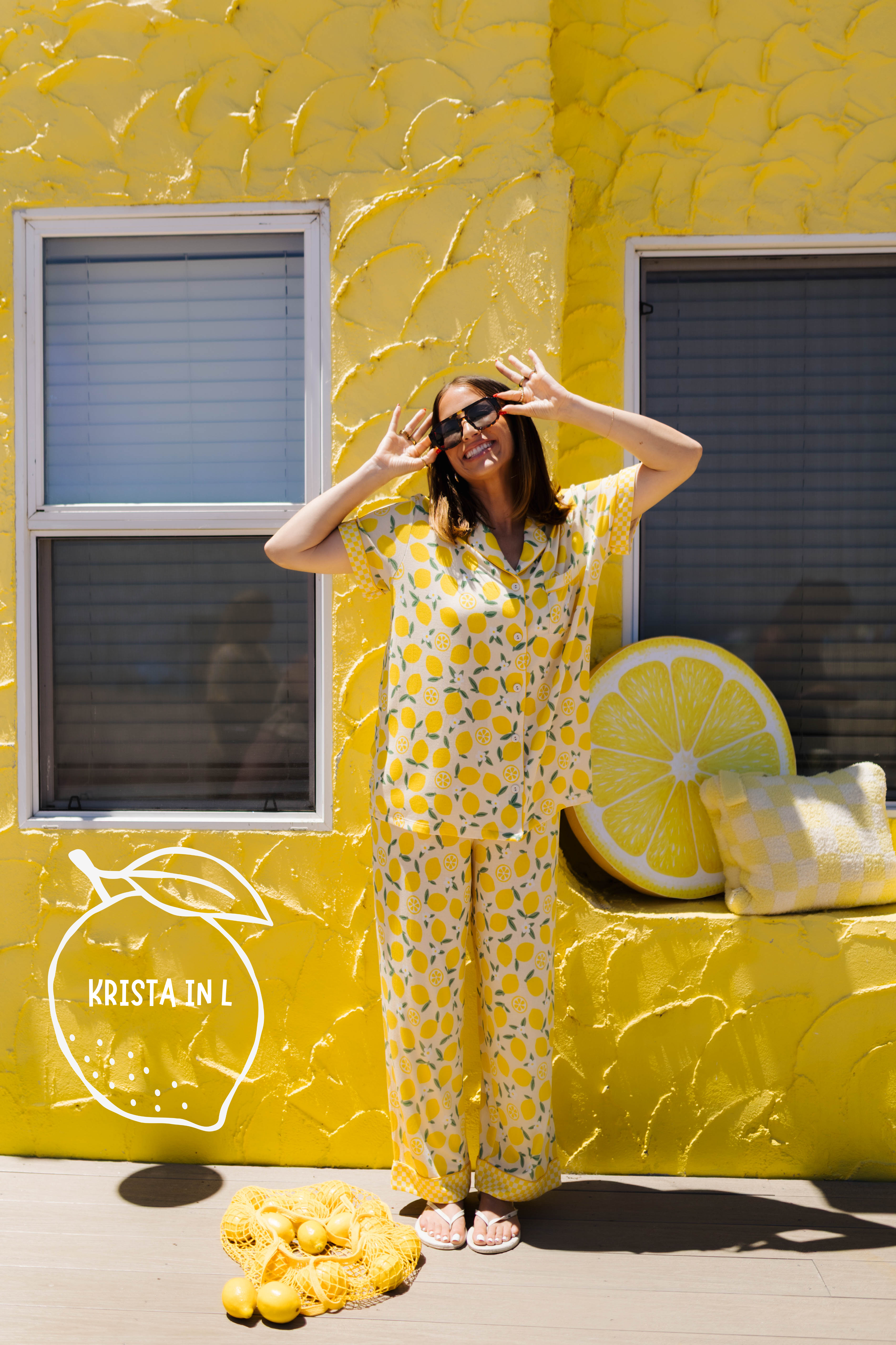 A woman wearing a yellow lemon-patterned pajama set stands in front of a bright yellow textured wall. The scene includes a large lemon slice prop on the wall, a pile of lemons on the ground, and a logo overlay reading 'Kristin L'.