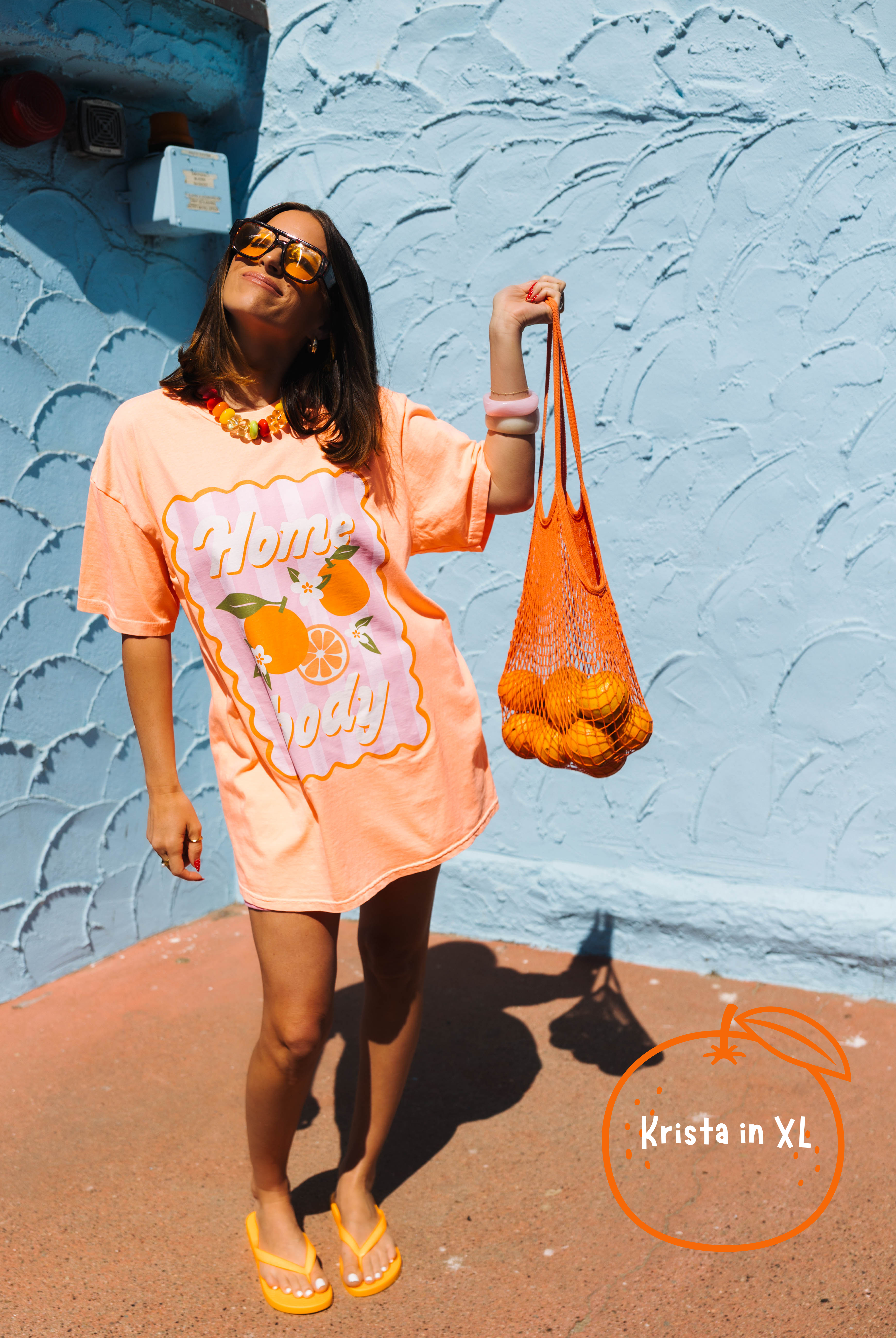A woman wearing an oversized peach-orange graphic t-shirt dress with 'Home Body' text and orange illustrations, holding a mesh bag of oranges against a textured light blue wall.
