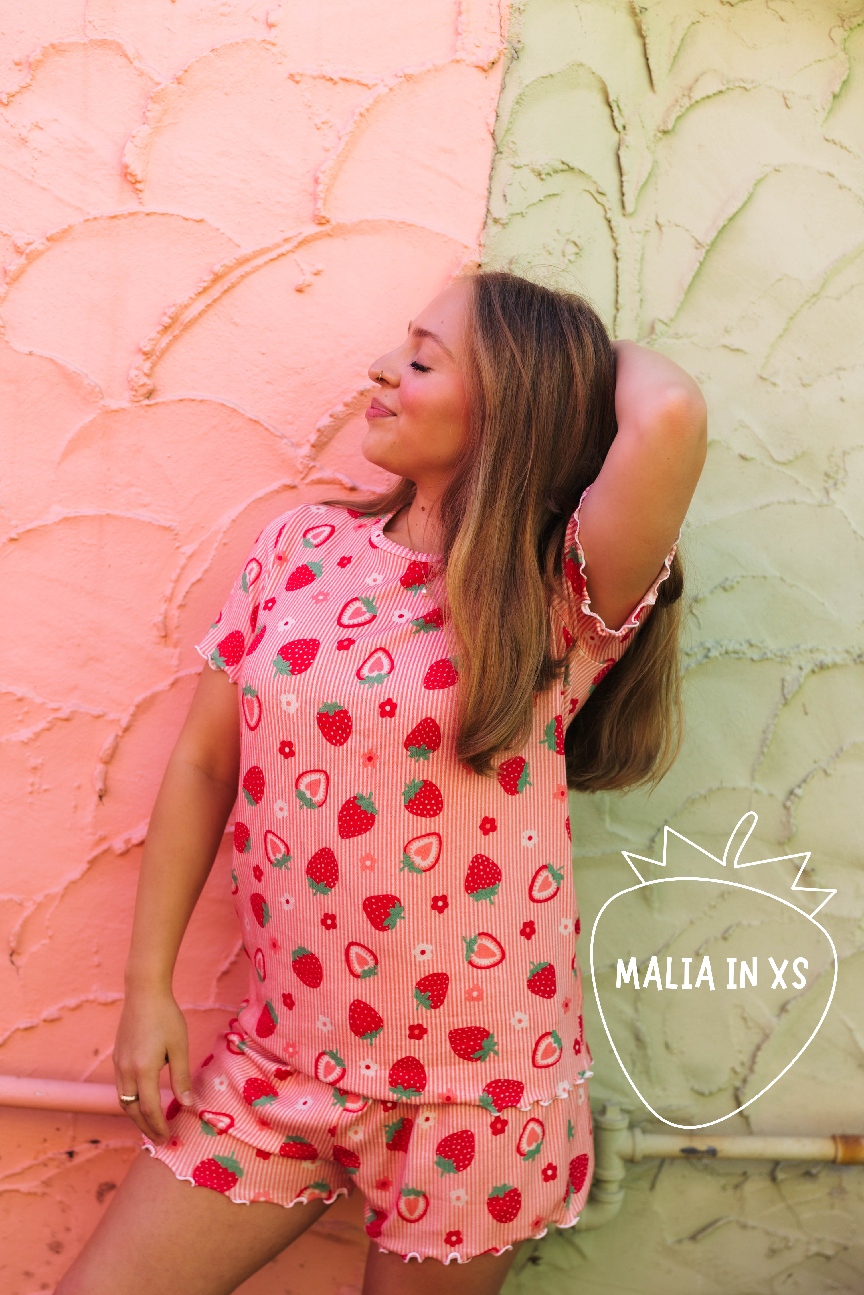 A woman wearing a pink strawberry-print pj set, posing against a split pink and green textured wall. A logo for 'MALIA IN XS' is visible in the bottom right corner.