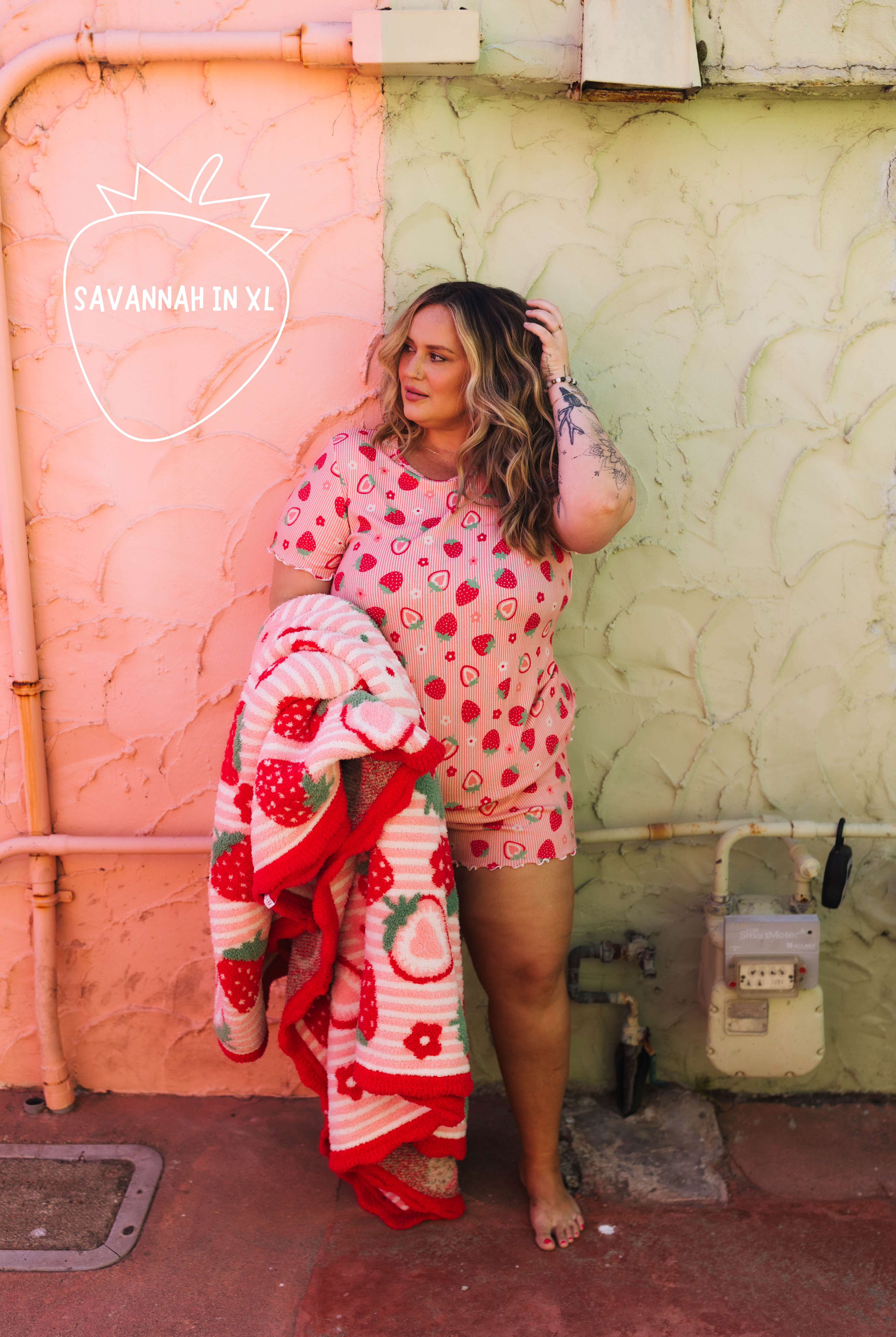 A woman stands outdoors against a split pink and green wall, wearing a pink strawberry-print short-sleeve top and matching shorts while holding a matching strawberry-patterned blanket. A white 'SAVANNAH IN XL' logo watermark is visible on the left side.