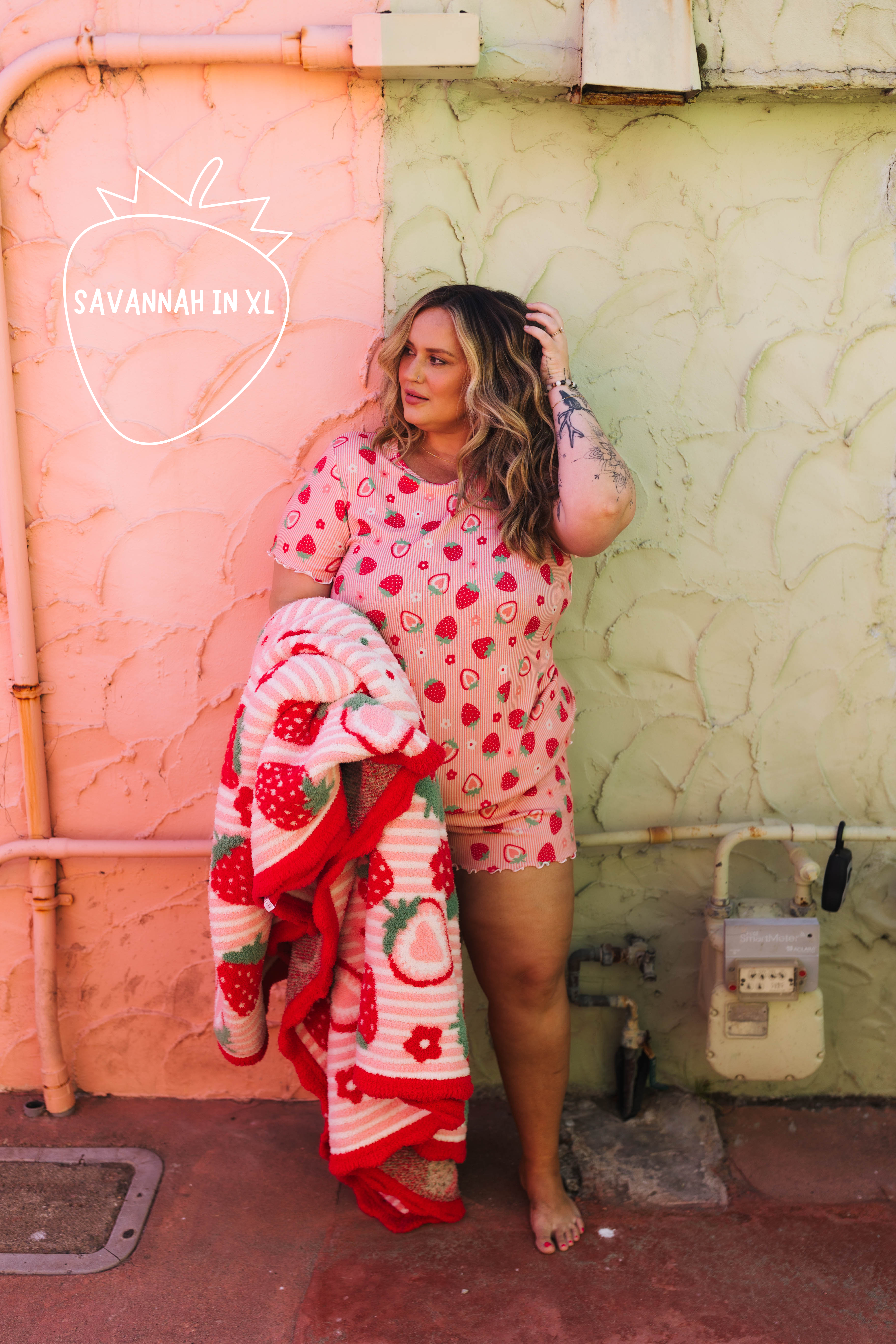 A woman stands outdoors against a split pink and green wall, wearing a pink strawberry-print short-sleeve top and matching shorts while holding a matching strawberry-patterned blanket. A white 'SAVANNAH IN XL' logo watermark is visible on the left side.