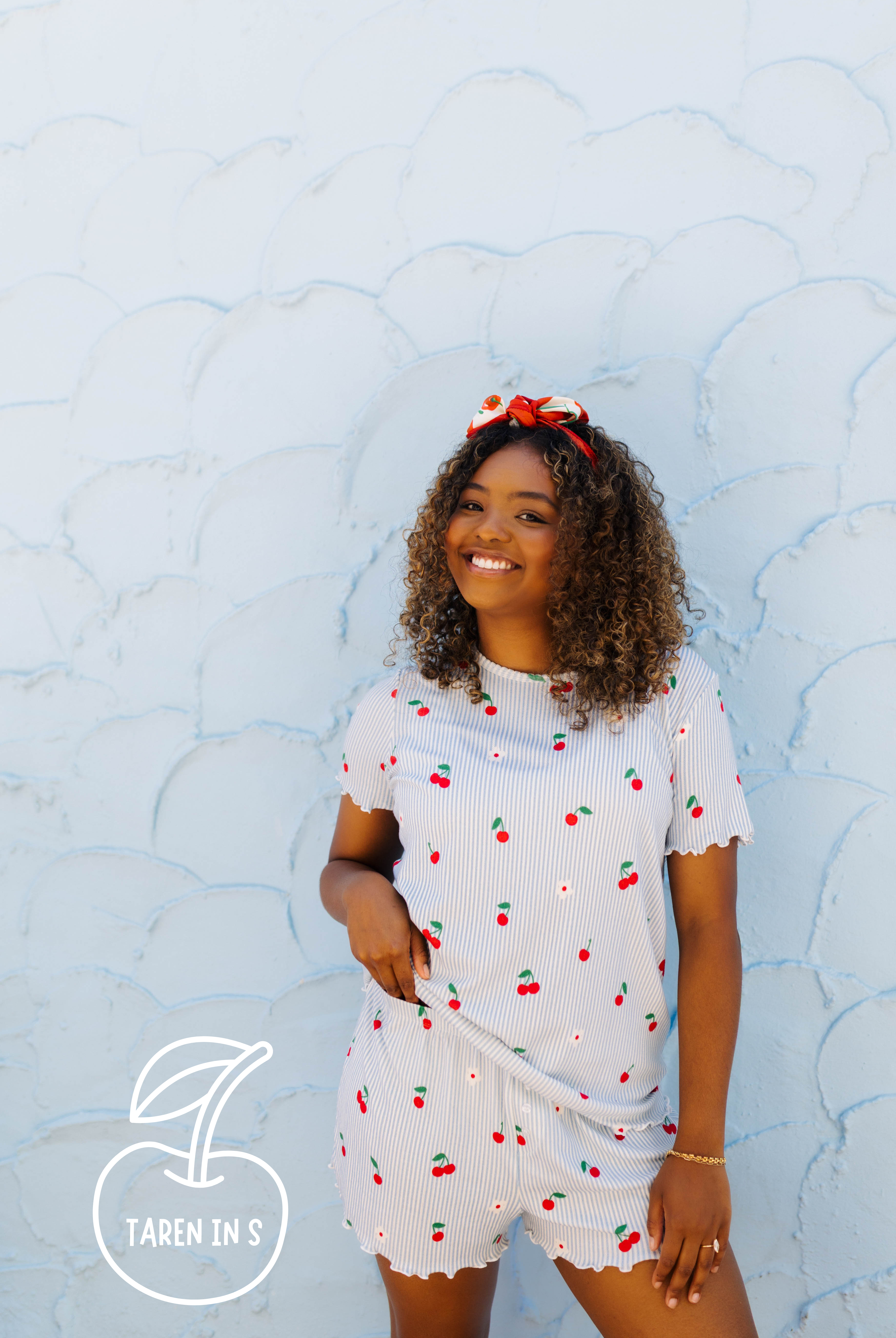A woman wearing a white ribbed short-sleeve pj set with a cherry print, standing against a light blue textured wall. She has a red bow in her hair, and a logo for 'TAREN IN S' is visible in the bottom-left corner.