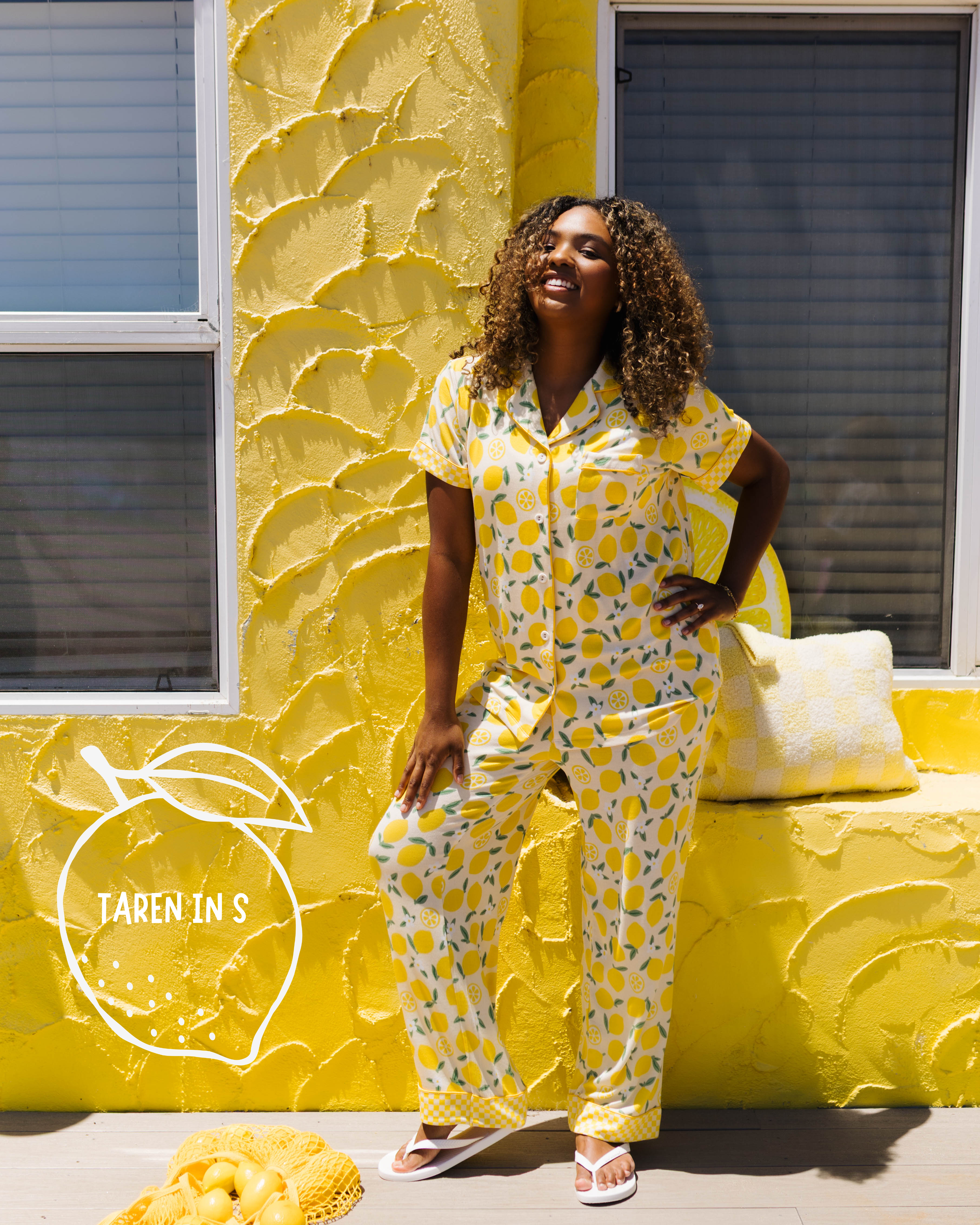 A woman modeling a yellow lemon-print pajama set stands in front of a textured yellow wall with windows. A pile of lemons and a small sign reading 'TAREN IN S' are on the ground nearby.