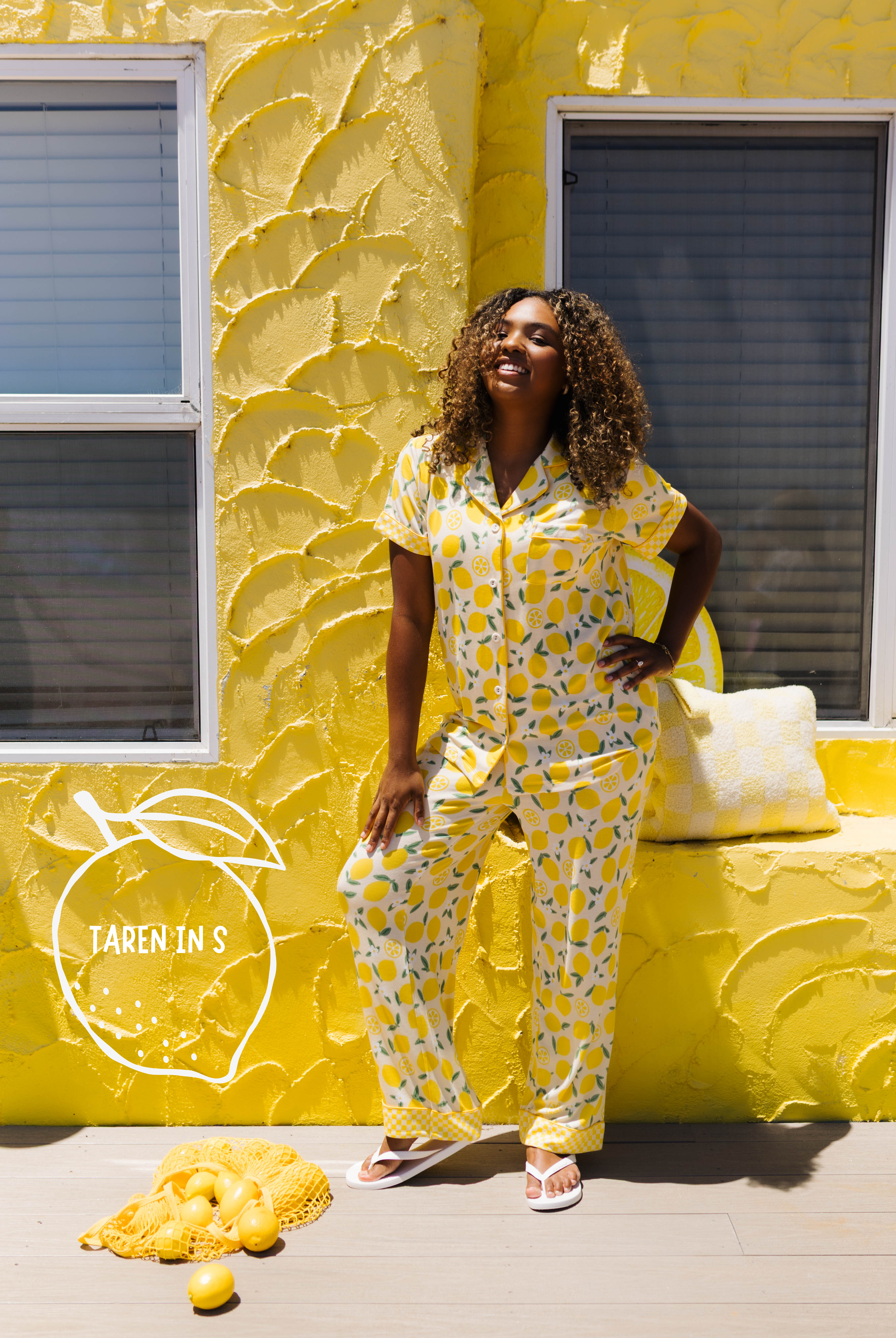 A woman modeling a yellow lemon-print pajama set stands in front of a textured yellow wall with windows. A pile of lemons and a small sign reading 'TAREN IN S' are on the ground nearby.