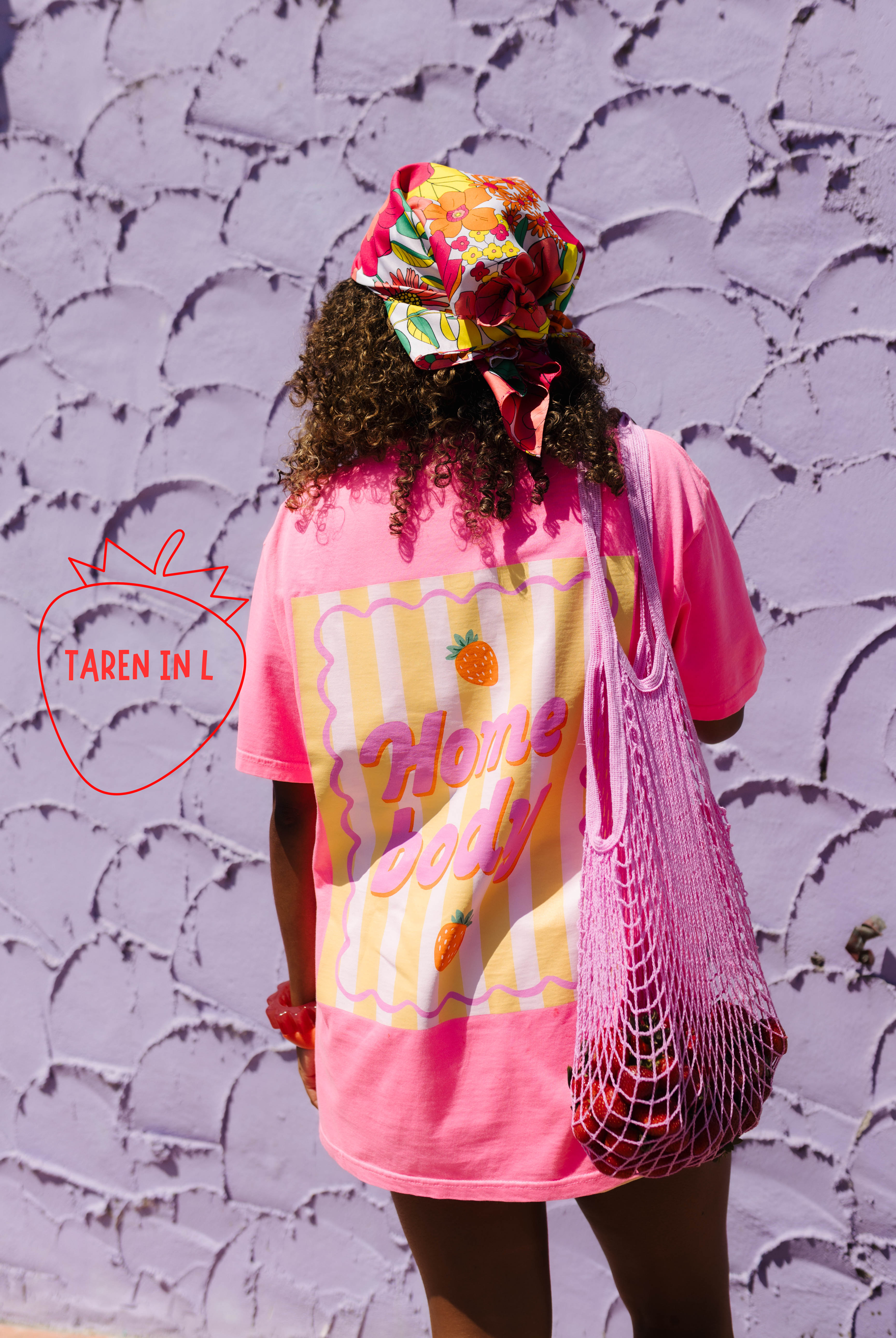 A person viewed from behind wearing a bright pink graphic T-shirt with a yellow and white retro wave design and the words 'homde body' on the back, paired with a colorful floral headwrap and a pink mesh tote bag, standing against a textured lavender wall.