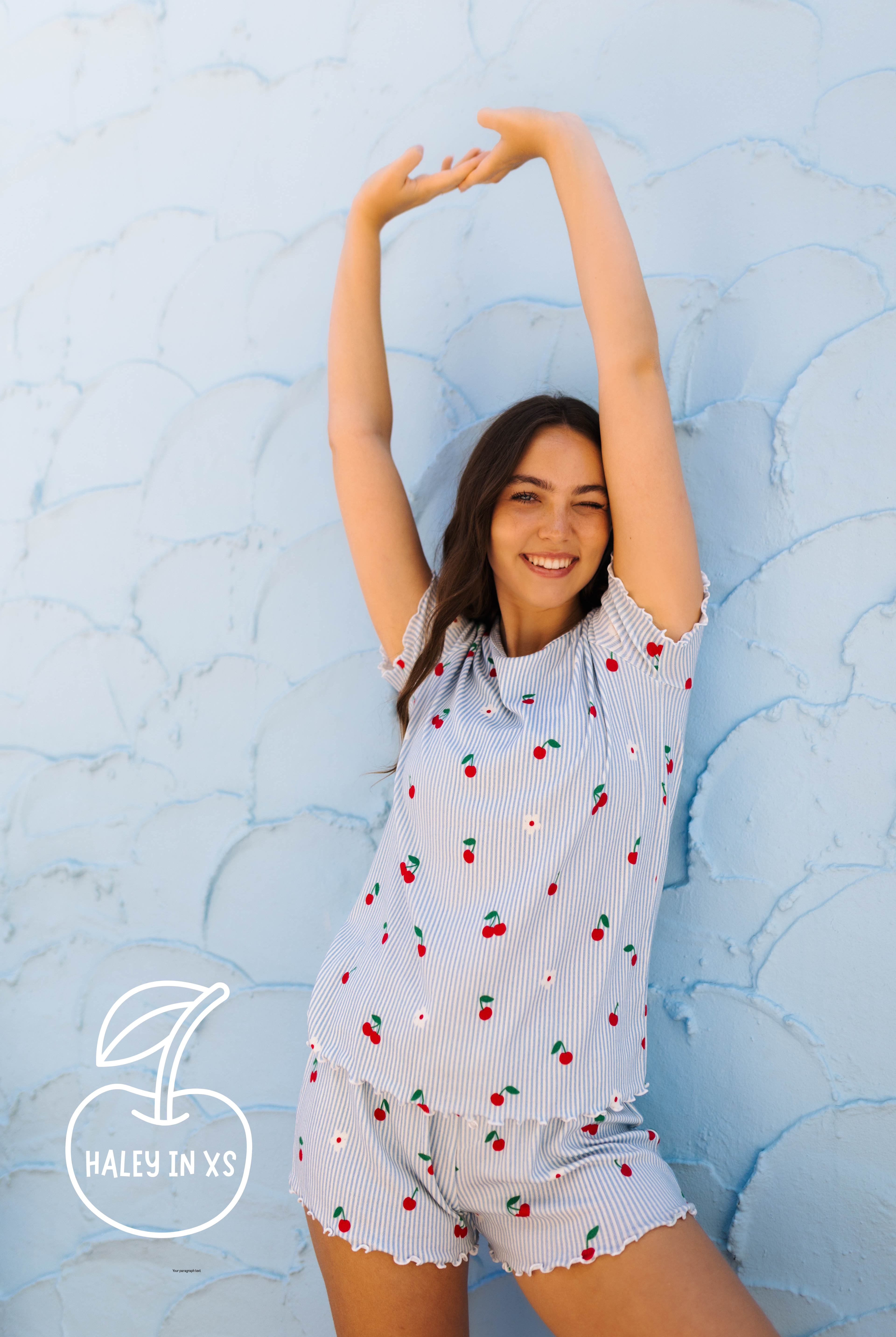 A woman wearing a light blue and white striped short-sleeve pajama set with a cherry print, posing against a textured light blue wall. A logo overlay in the bottom left reads 'HALEY IN XS'.