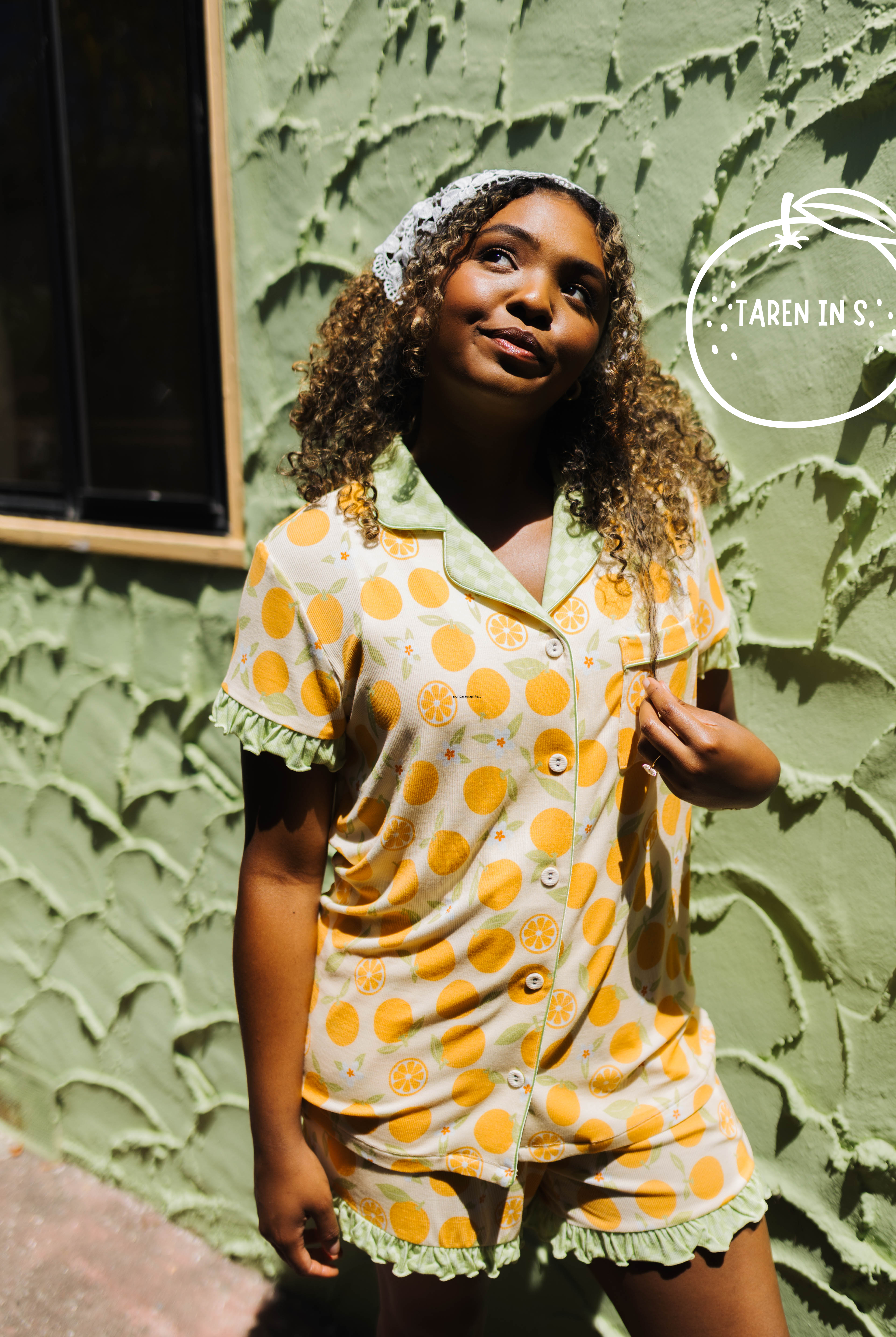 A woman wearing a two-piece pajama set with a orange citrus pattern on a cream background, standing against a textured green stucco wall. She is wearing a matching patterned headband.
