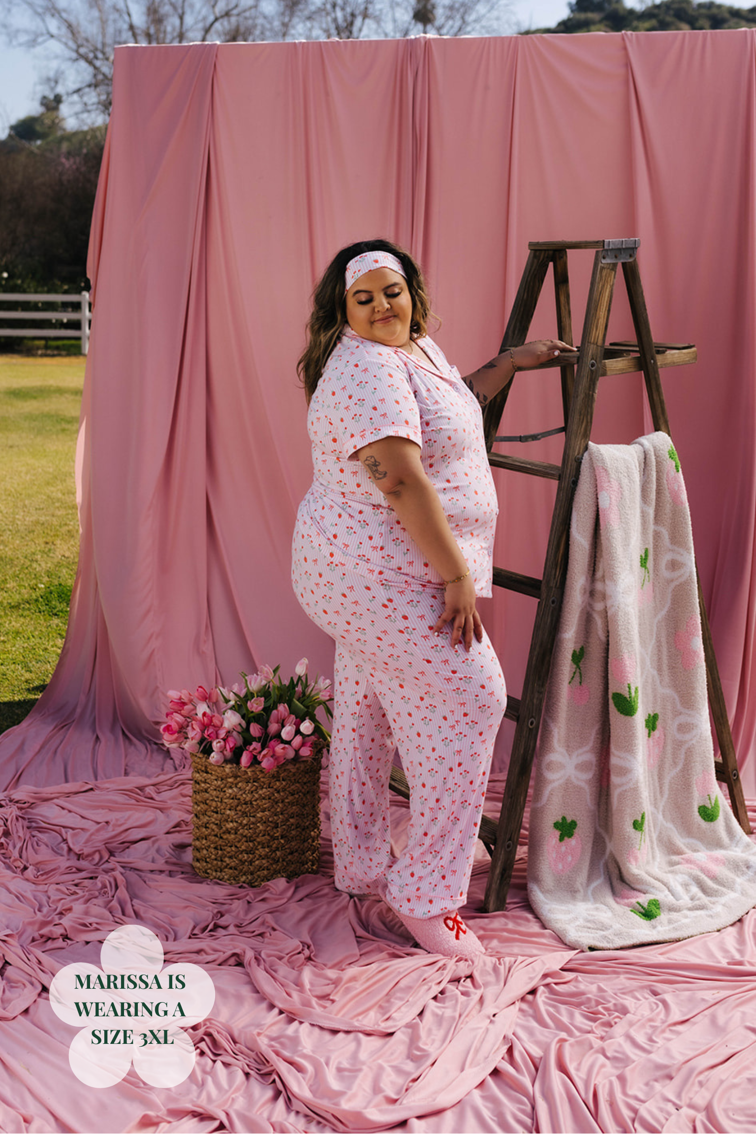 Woman in a floral outfit standing next to a wooden ladder and pink fabric backdrop.