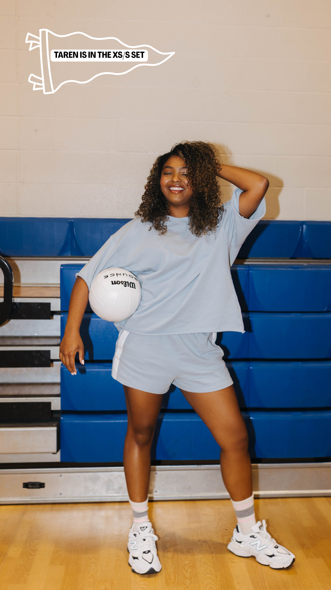 A woman posing in a gym wearing a light blue oversized t-shirt and matching shorts, holding a white ball. 