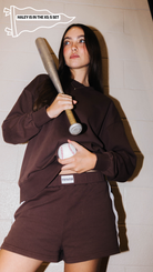 A woman modeling a dark sweatshirt and matching shorts, holding a baseball bat and a baseball against a white brick wall. A text overlay in the top left reads 'HALEY IS IN THE XS-S SET'.