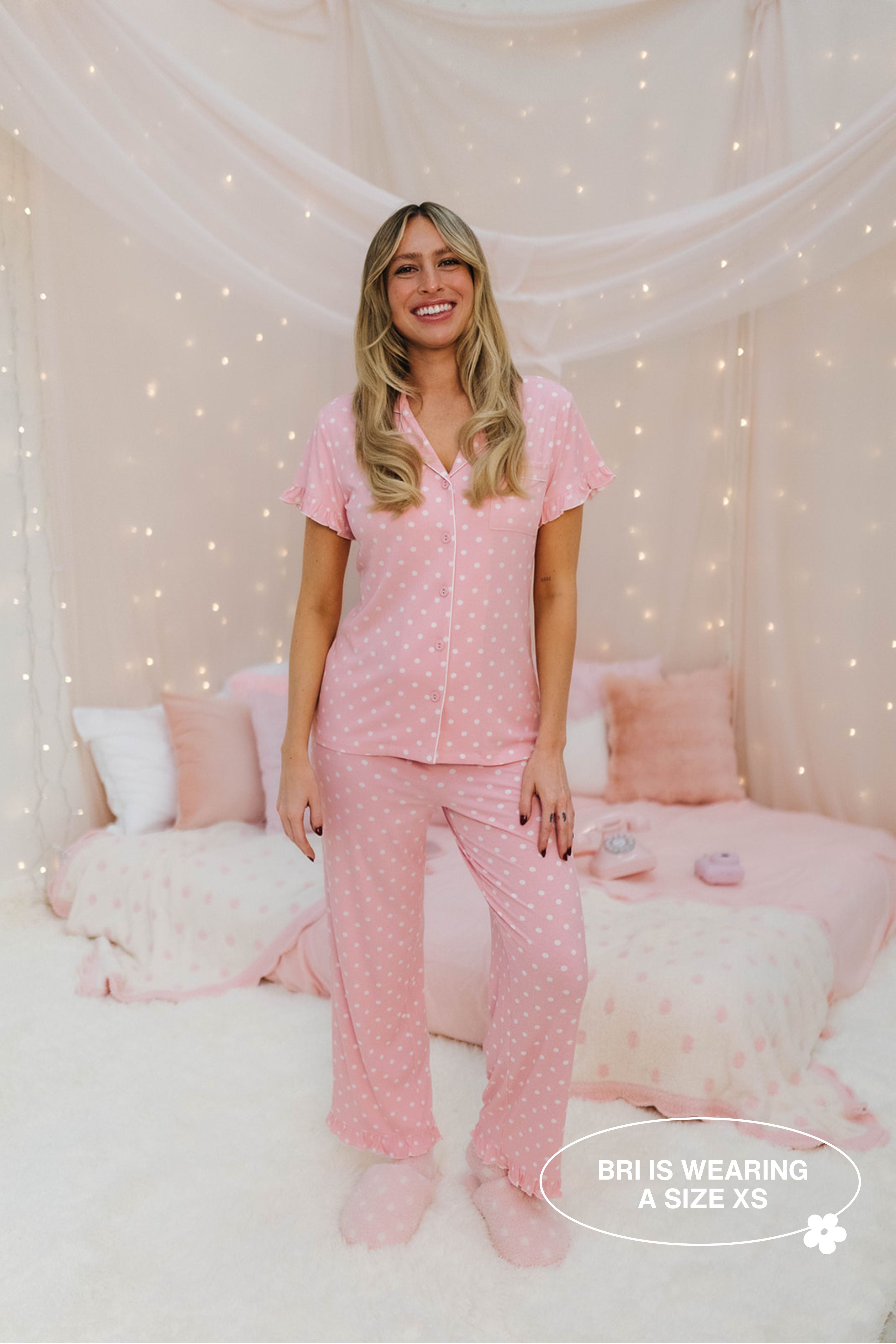 Woman wearing pink pajamas sitting on a bed with decorative pillows and fairy lights.